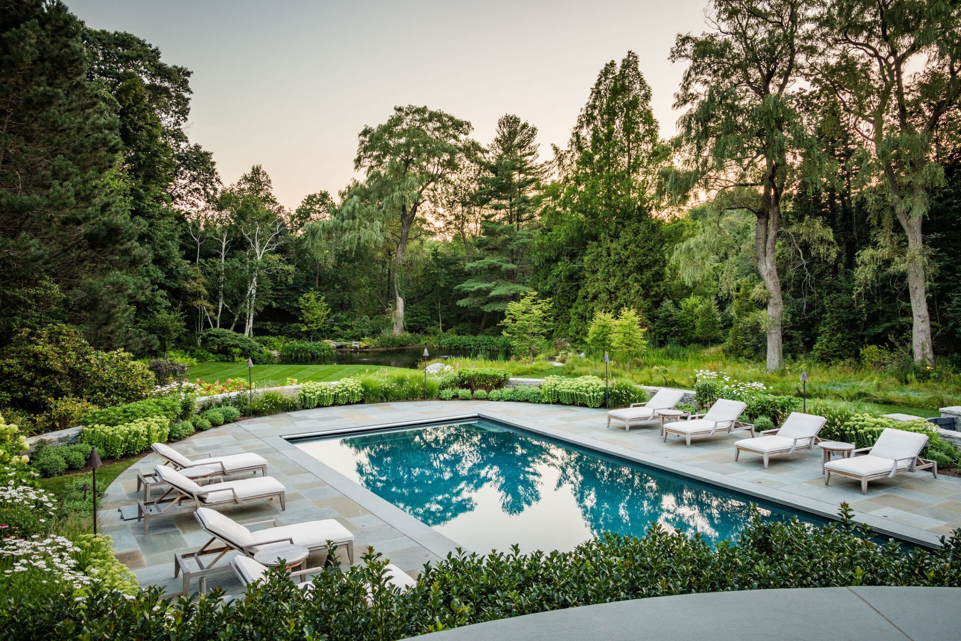 An outdoor swimming pool surrounded by lounge chairs, neatly landscaped gardens, and lush trees reflects the tranquil dusk sky in a serene setting.
