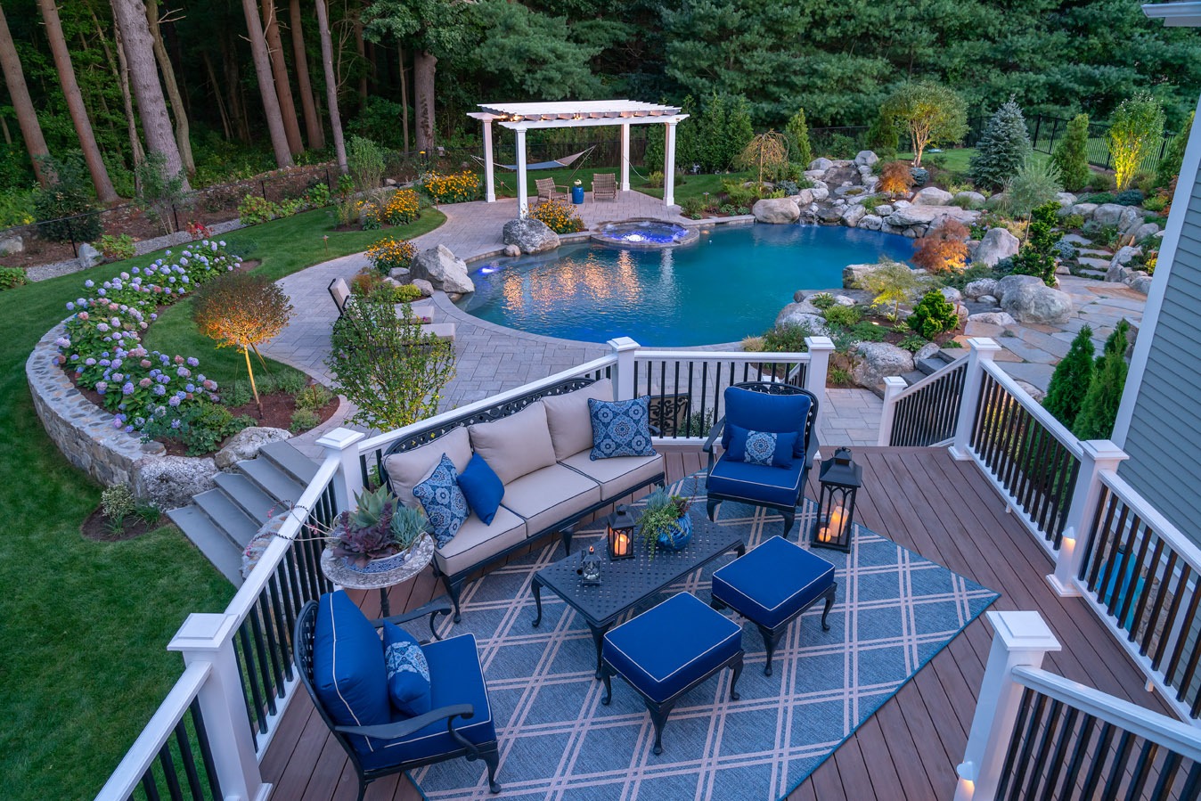 A beautifully landscaped backyard with a pool, pergola, and outdoor seating on a deck during twilight. Trees and lush plants surround the serene area.