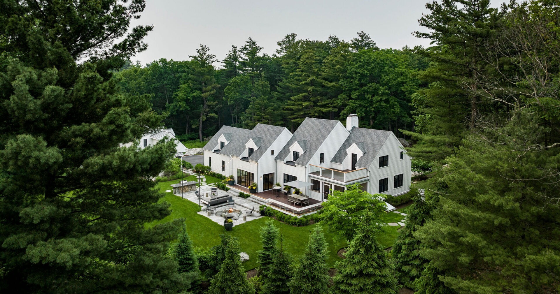 A large, luxurious white house with grey roofs, nestled among lush green trees, boasts landscaped gardens and a spacious outdoor entertaining area.