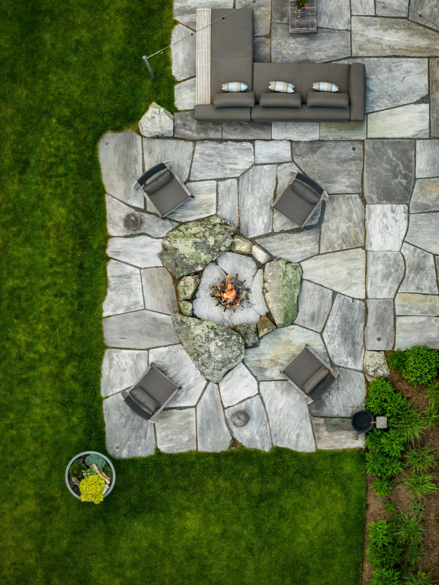 An aerial view of an outdoor stone patio with a fire pit in the center surrounded by chairs, adjacent to a neatly trimmed lawn.