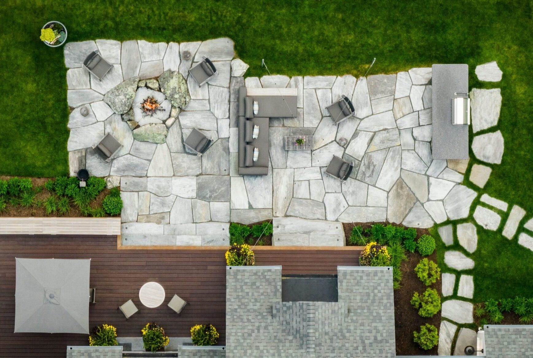 An aerial view of a landscaped backyard featuring a stone patio with a fire pit, outdoor furniture, green lawn, and structured garden beds.