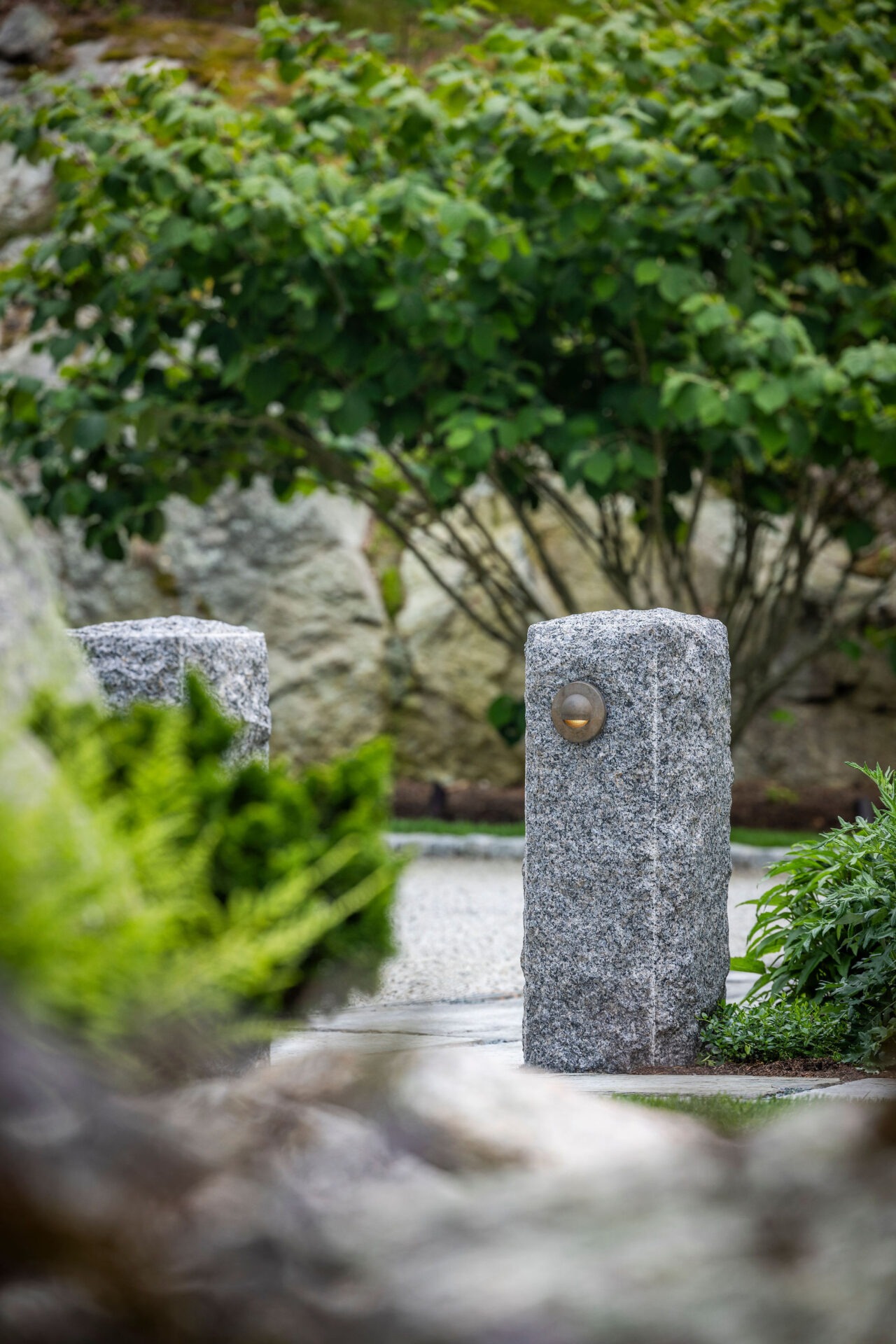 This image features two granite posts with round features, possibly lights, set in a tranquil garden with lush greenery and a pebbled path.