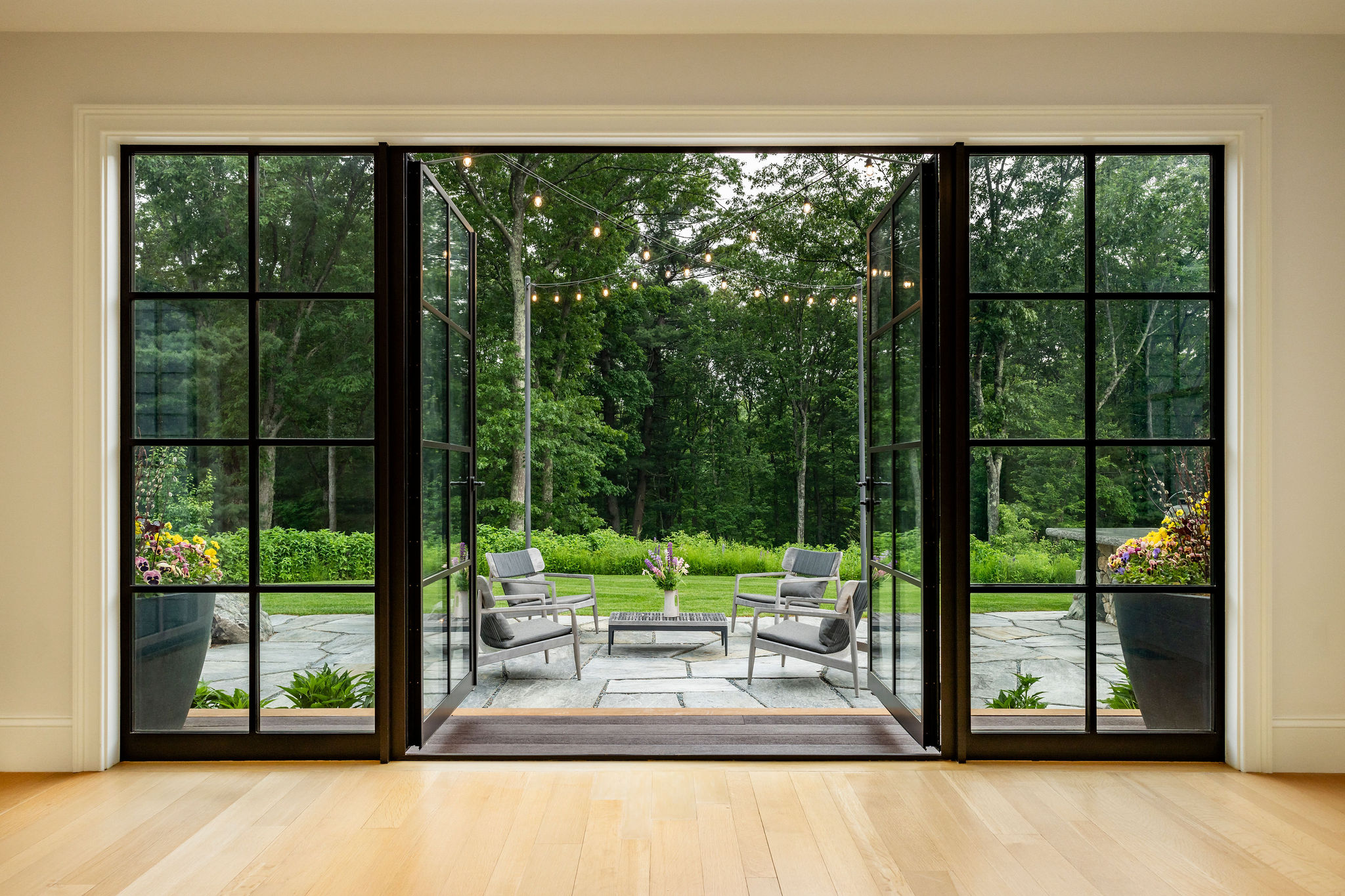 A spacious, well-lit room opens to a patio with furniture, string lights, and a lush green background through large black-framed glass doors.