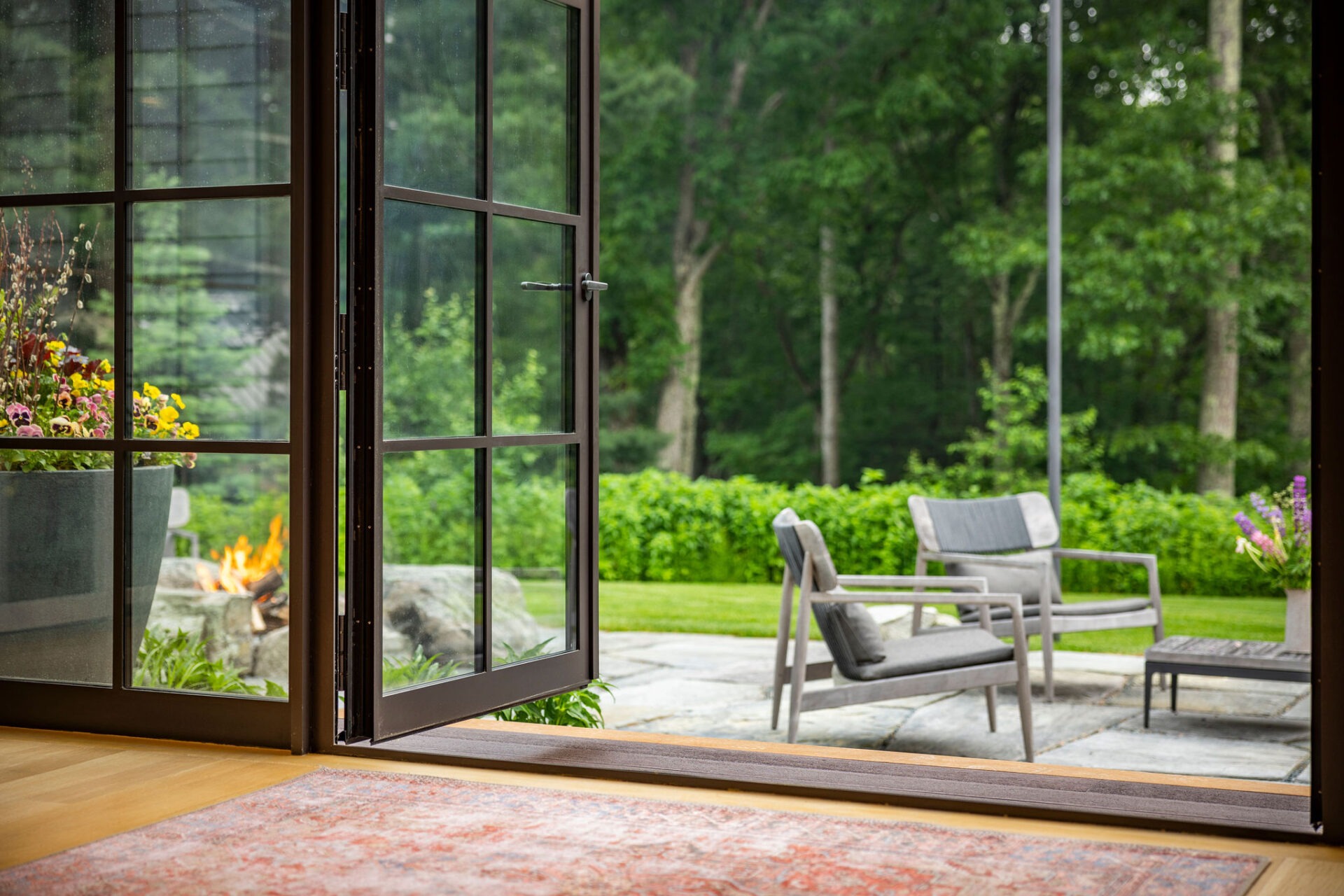 This image shows a peaceful outdoor patio view through an open glass door. Comfortable furniture, a lit fire pit, and lush greenery create a serene scene.