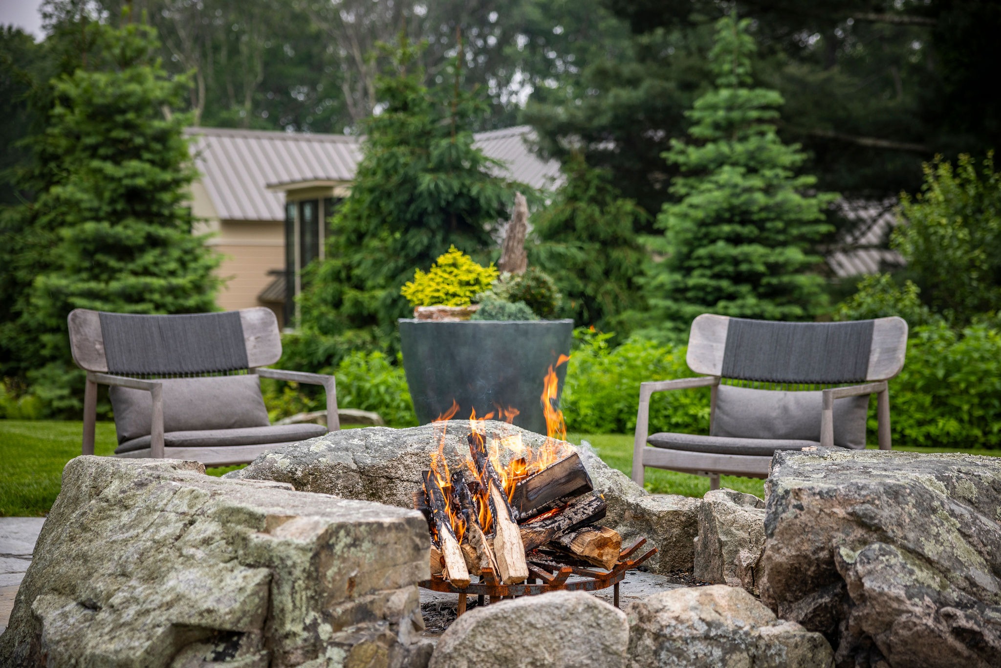 An outdoor fire pit with flames, surrounded by large rocks, with two armchairs facing it, in a lush garden setting with trees and a shrub pot.