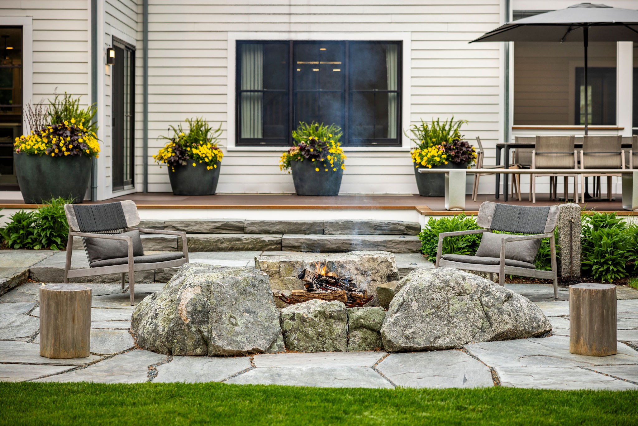 An outdoor patio with chairs and a fire pit. Large planters with yellow flowers flank a house. A dining area with an umbrella is in the background.