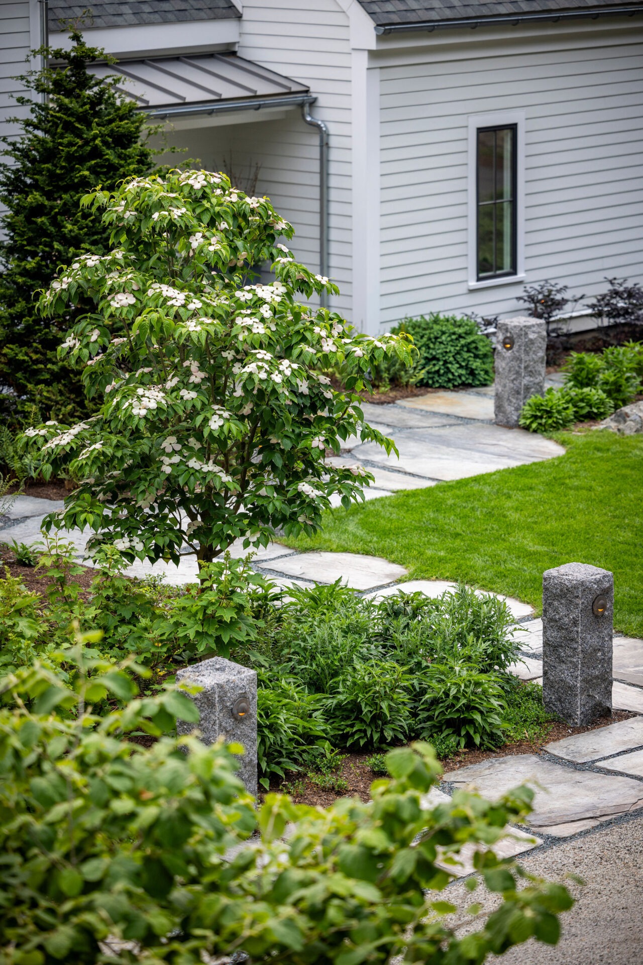 This image shows a landscaped garden with a flowering bush, green shrubs, a stone pathway, and granite posts by a grey house.