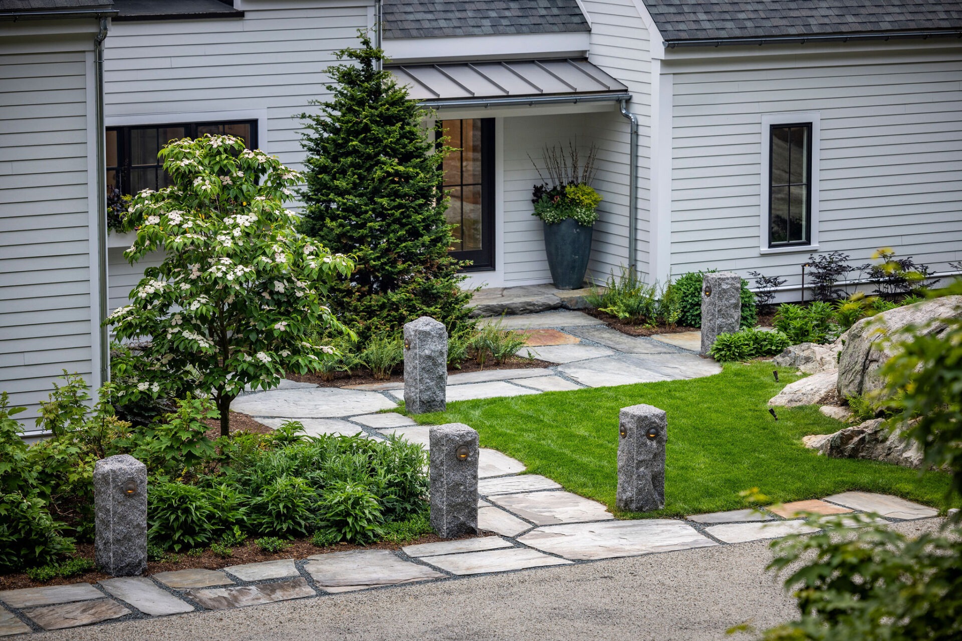 A well-manicured garden with a stone path, green lawn, and decorative granite posts leading to a gray house with white trim under a cloudy sky.