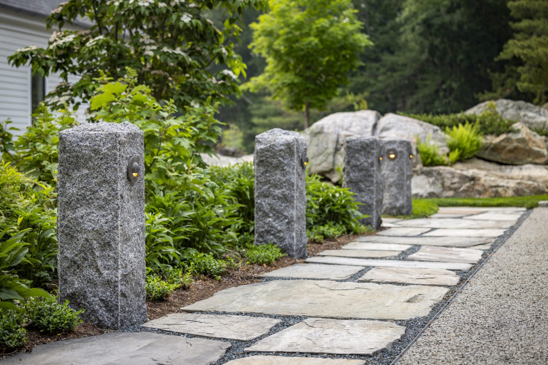 A landscaped pathway lined with gray stone posts featuring built-in lights. Green shrubbery and a large rock formation create a tranquil, natural setting.