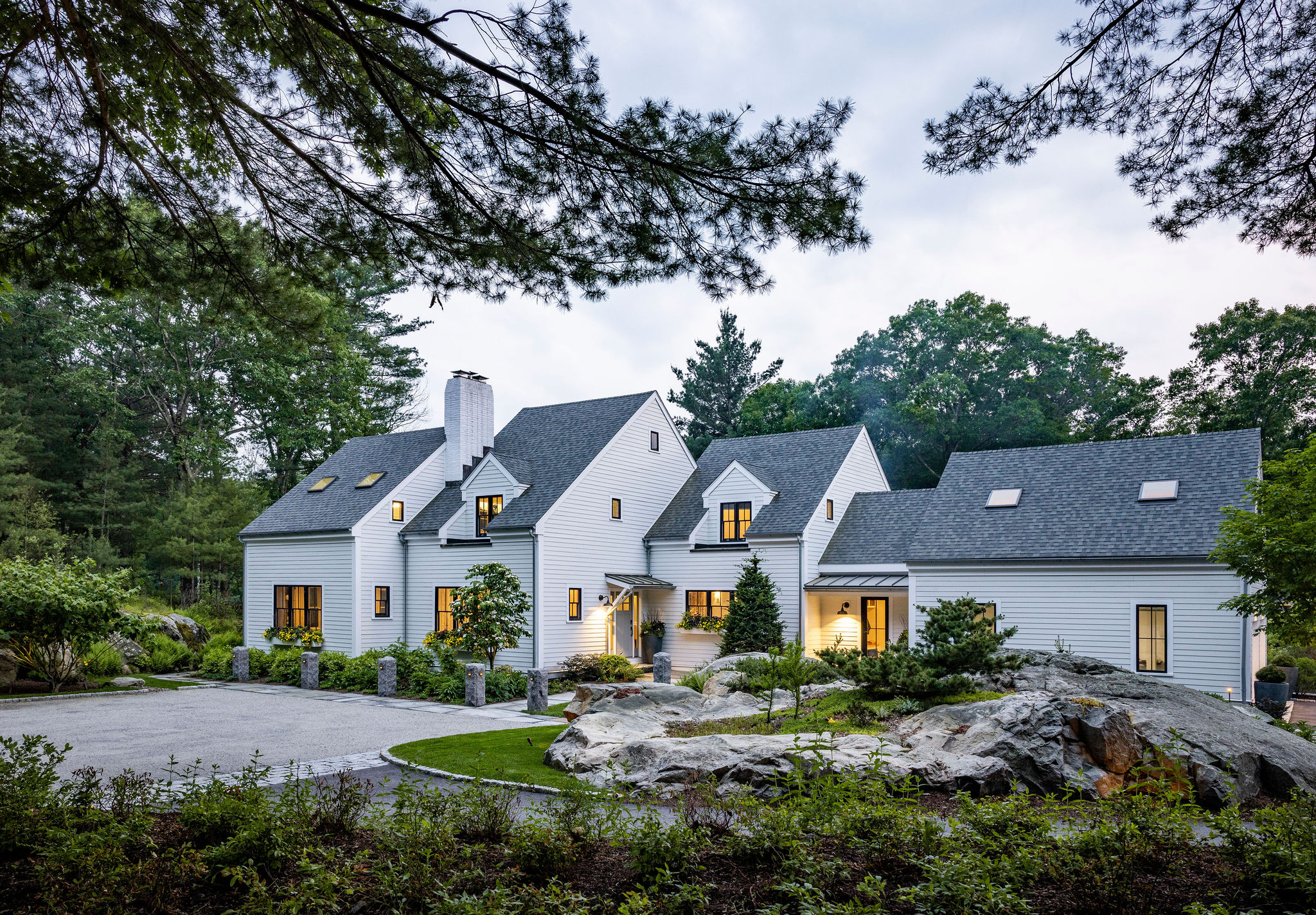 The image shows a large, white two-story house with a gabled roof, surrounded by trees, landscaping, and a circular driveway at dusk.