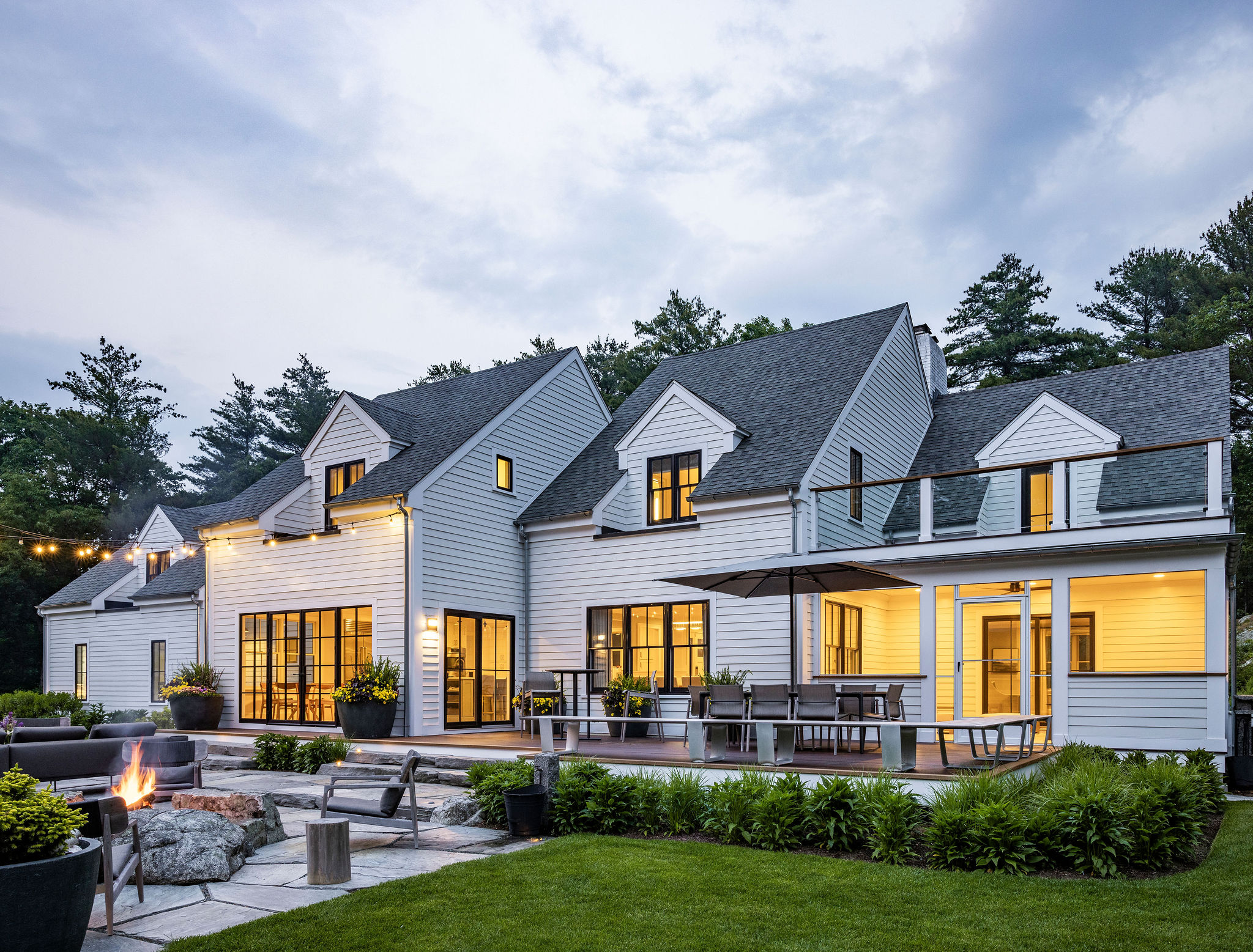 A modern two-story house with white siding during dusk. Warm interior lights glow, a fire pit burns outside, and string lights add ambiance.