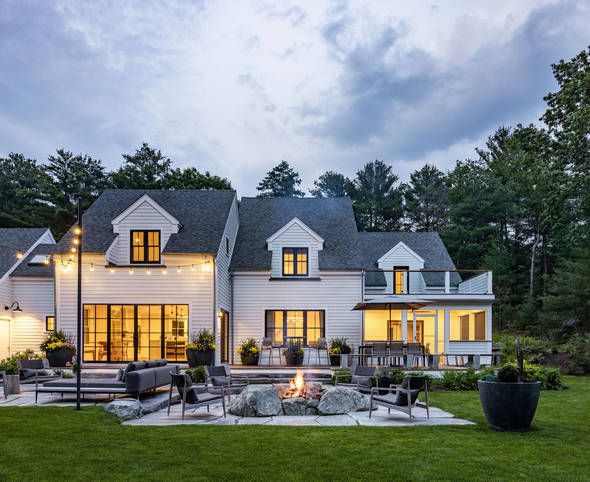 The image shows a modern two-story house with white siding, illuminated by interior lighting and exterior string lights, featuring a backyard with a fire pit and seating.