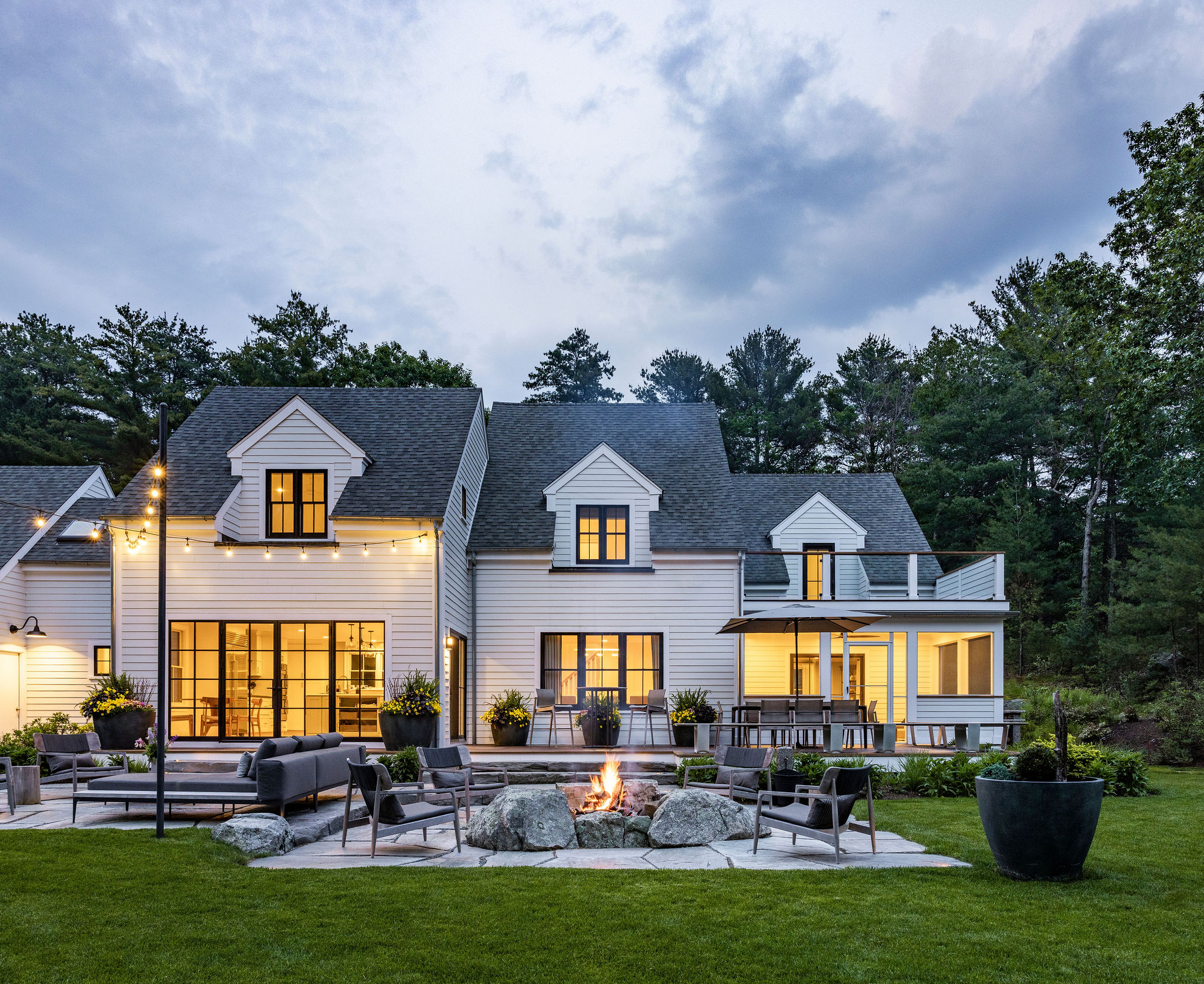 The image shows a modern two-story house with white siding, illuminated by interior lighting and exterior string lights, featuring a backyard with a fire pit and seating.