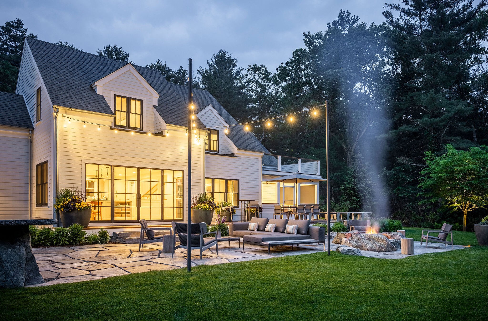 This image shows a modern two-story home at dusk, with outdoor lighting, a fire pit, lounge furniture, and a lush lawn surrounded by trees.