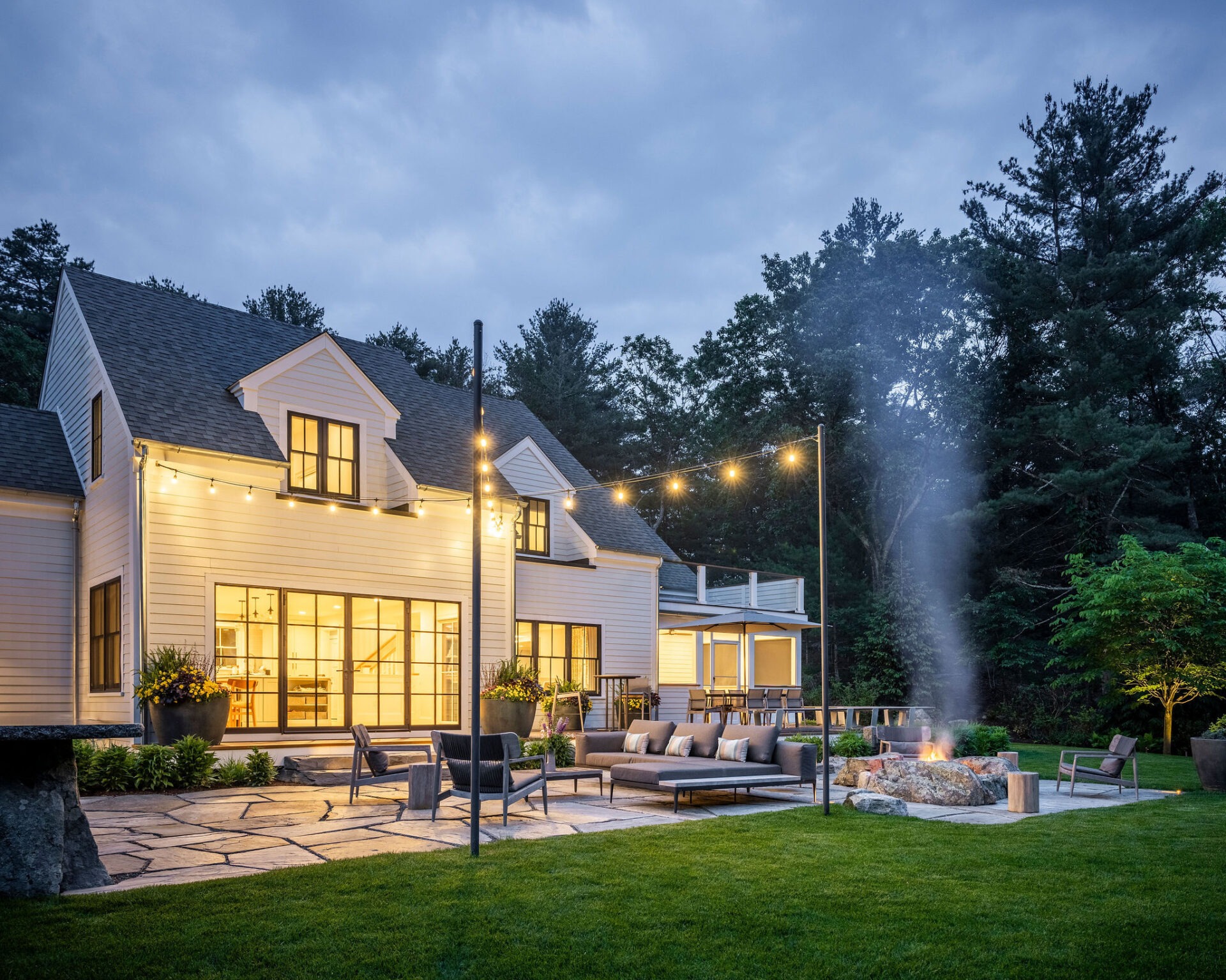 A dusk view of a cozy, illuminated backyard featuring an elegant home, strung lights, outdoor seating, a fire pit, well-maintained lawn, and trees.