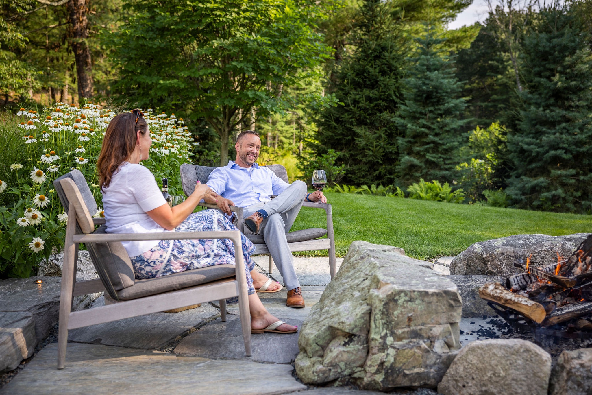 Two people are relaxing by a fire pit in a lush garden, with one holding a wine glass, amidst a backdrop of greenery and flowers.