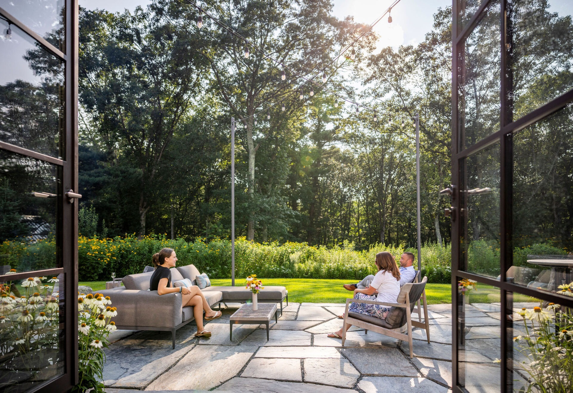 Three people relax on patio furniture in a lush garden with trees, flowers, and string lights, viewed from an open glass door.
