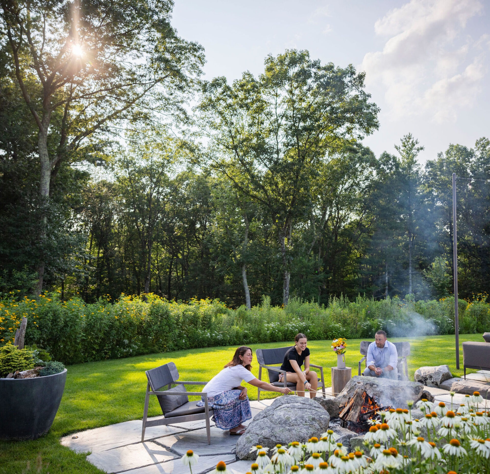 Three people are sitting in a backyard with a fire pit, surrounded by lush greenery, under a clear sky with sunlight streaming through the trees.