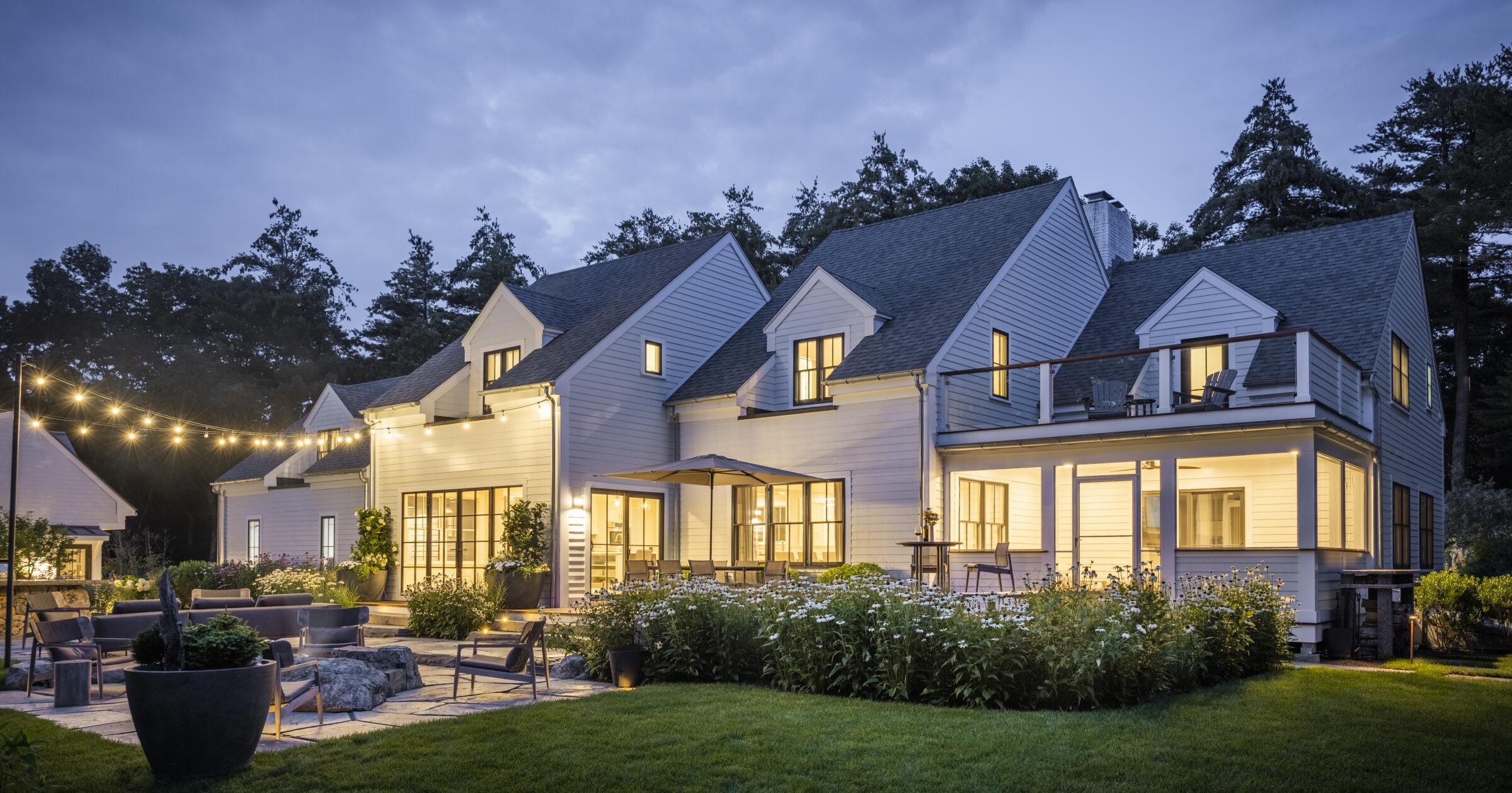A spacious two-story house at dusk with exterior lights on, featuring a patio with outdoor furniture, string lights, and a well-manicured lawn.