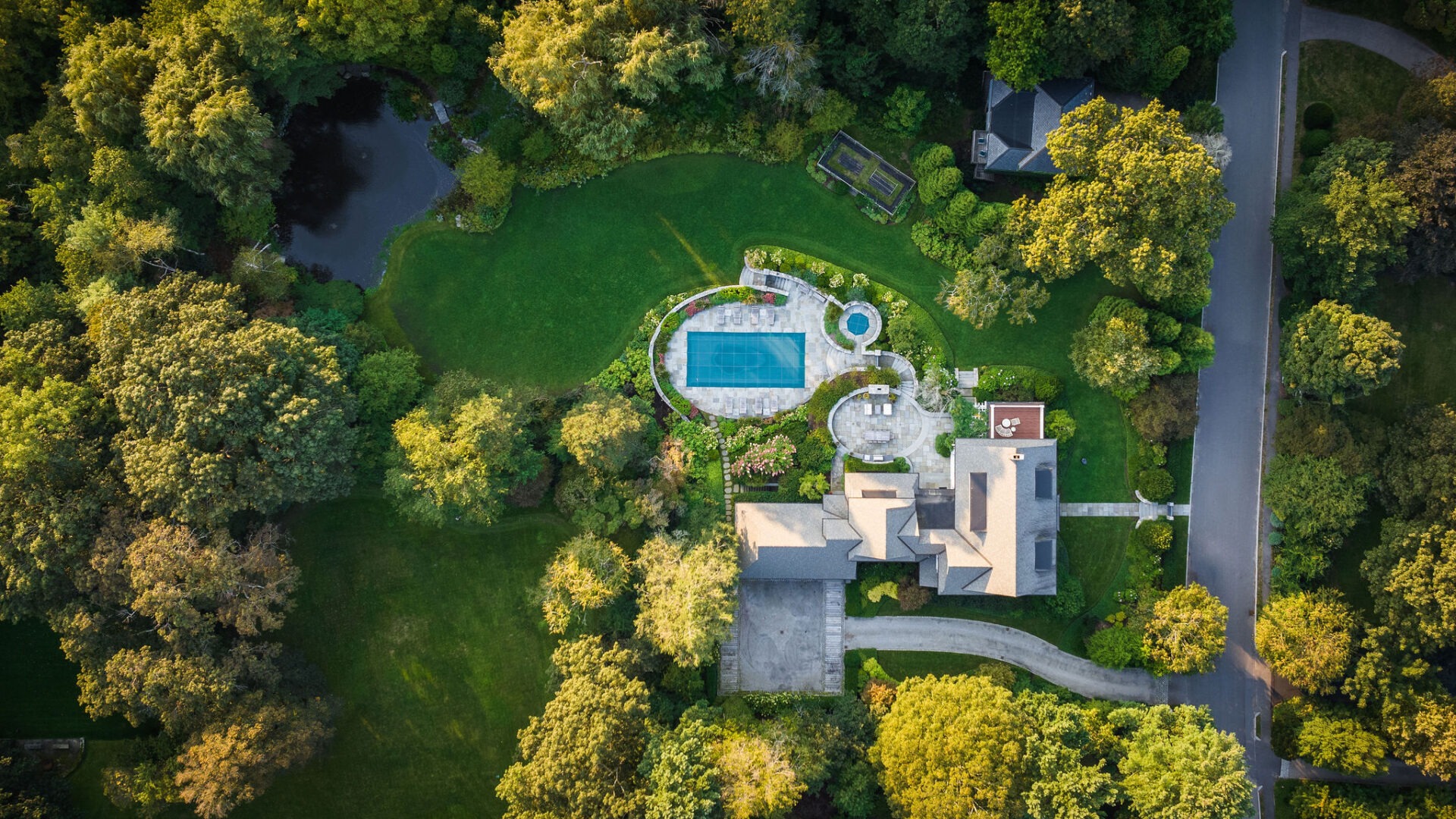 An aerial view of a luxurious estate with a large swimming pool, manicured gardens, surrounded by lush trees, and flanked by a curving driveway.