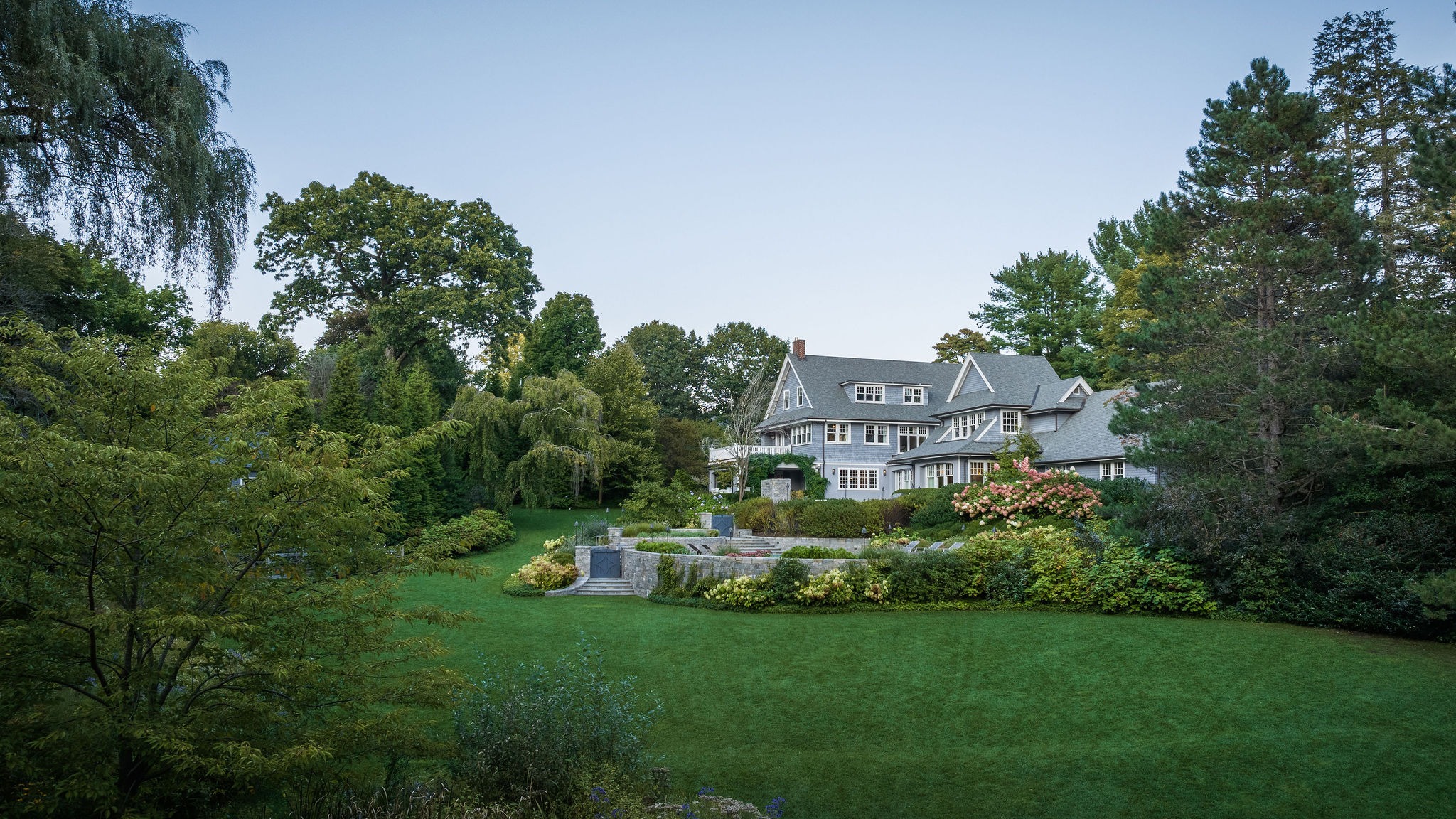 The image features a large, elegant house with gray siding surrounded by manicured gardens, lush trees, and a well-kept green lawn.