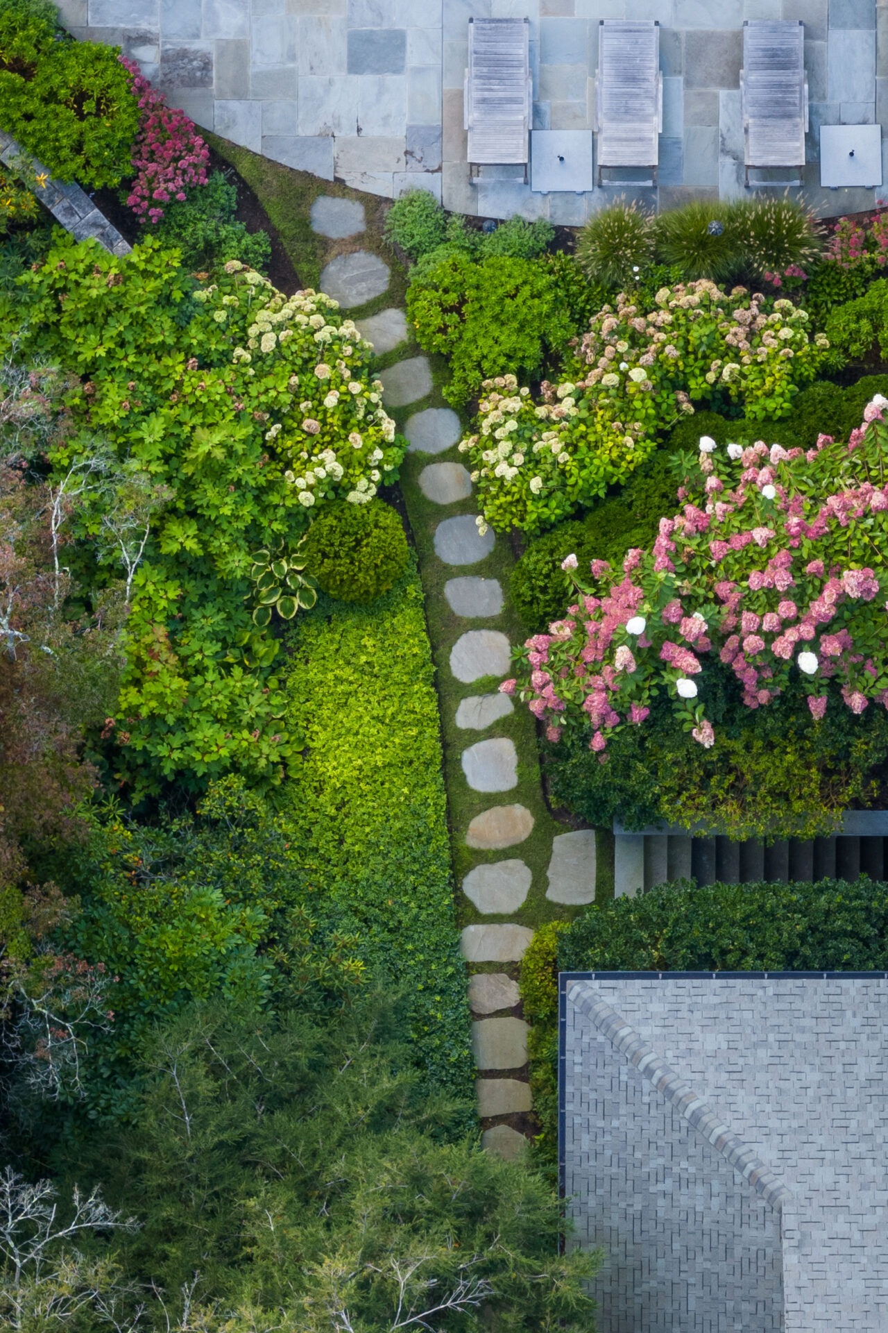 This aerial image shows a neatly arranged garden with a stone path leading through vibrant greenery and colorful flowering hydrangeas adjacent to a structure.