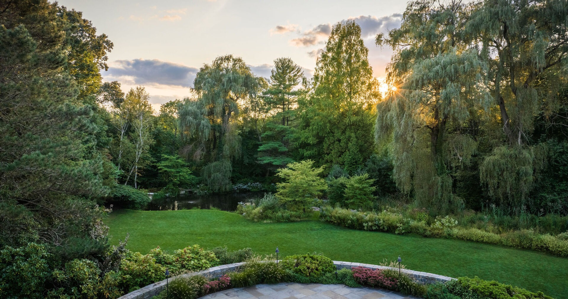 A serene garden at sunset with a pond, lush greenery, weeping willows, and a sun-kissed sky. A stone edge hints at a terrace in the foreground.