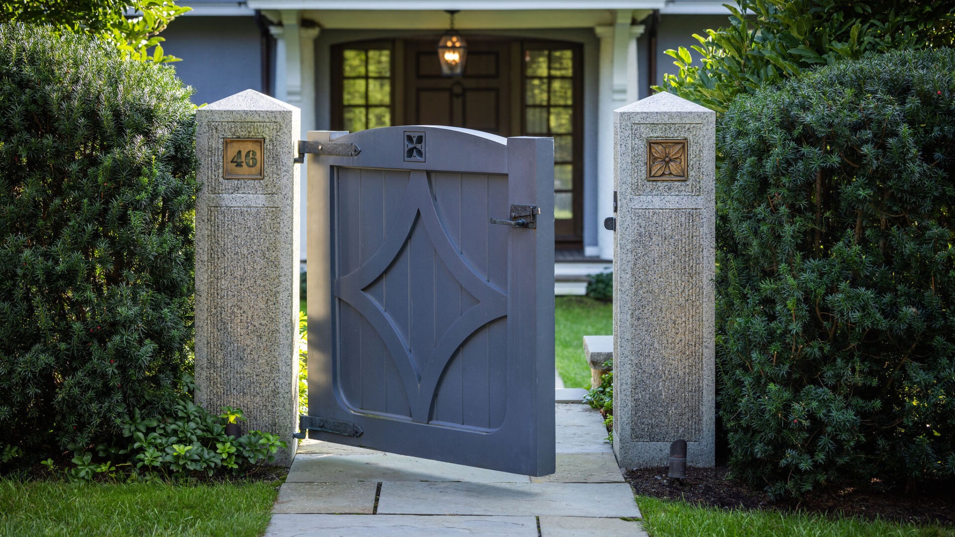 An elegant grey wooden gate with ornate patterns stands open between two stone pillars marked with the number '46', leading to a residential entrance.