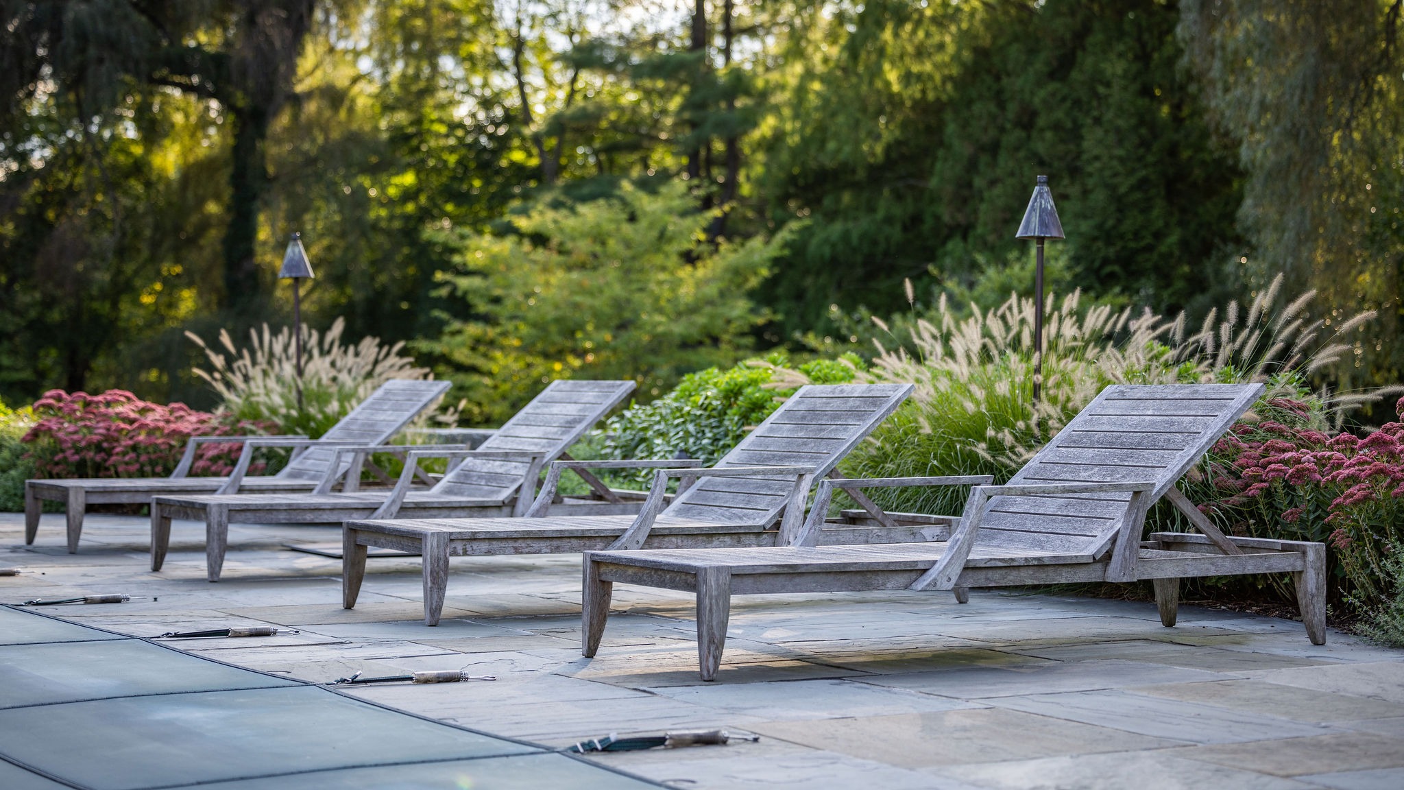 A serene outdoor setting with five weathered wooden lounge chairs arranged on a stone patio, surrounded by lush greenery and ornamental grasses under soft lighting.