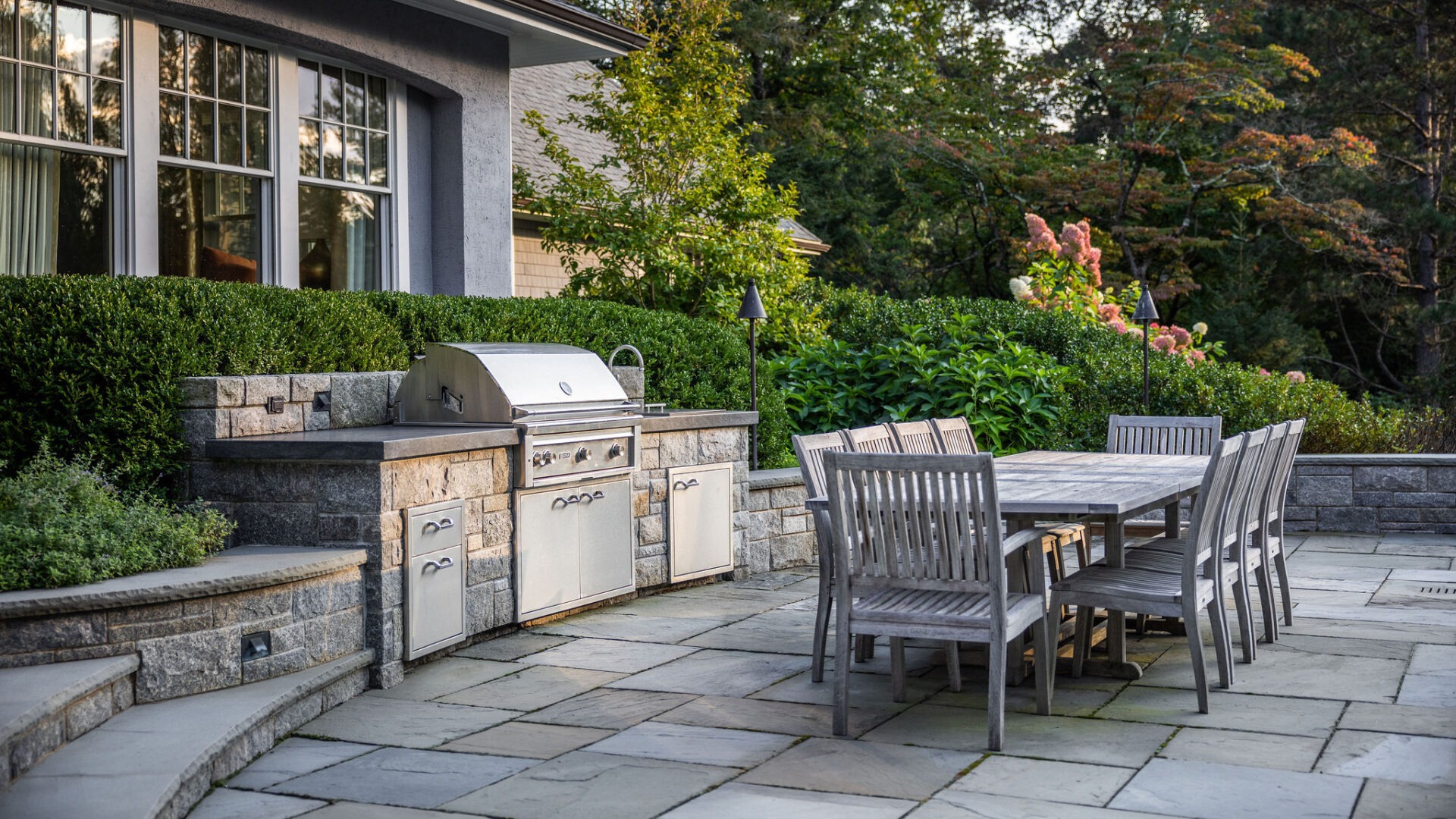 An outdoor patio featuring a stone-built kitchen area with a grill, surrounded by lush greenery, next to a wooden dining set for entertainment.