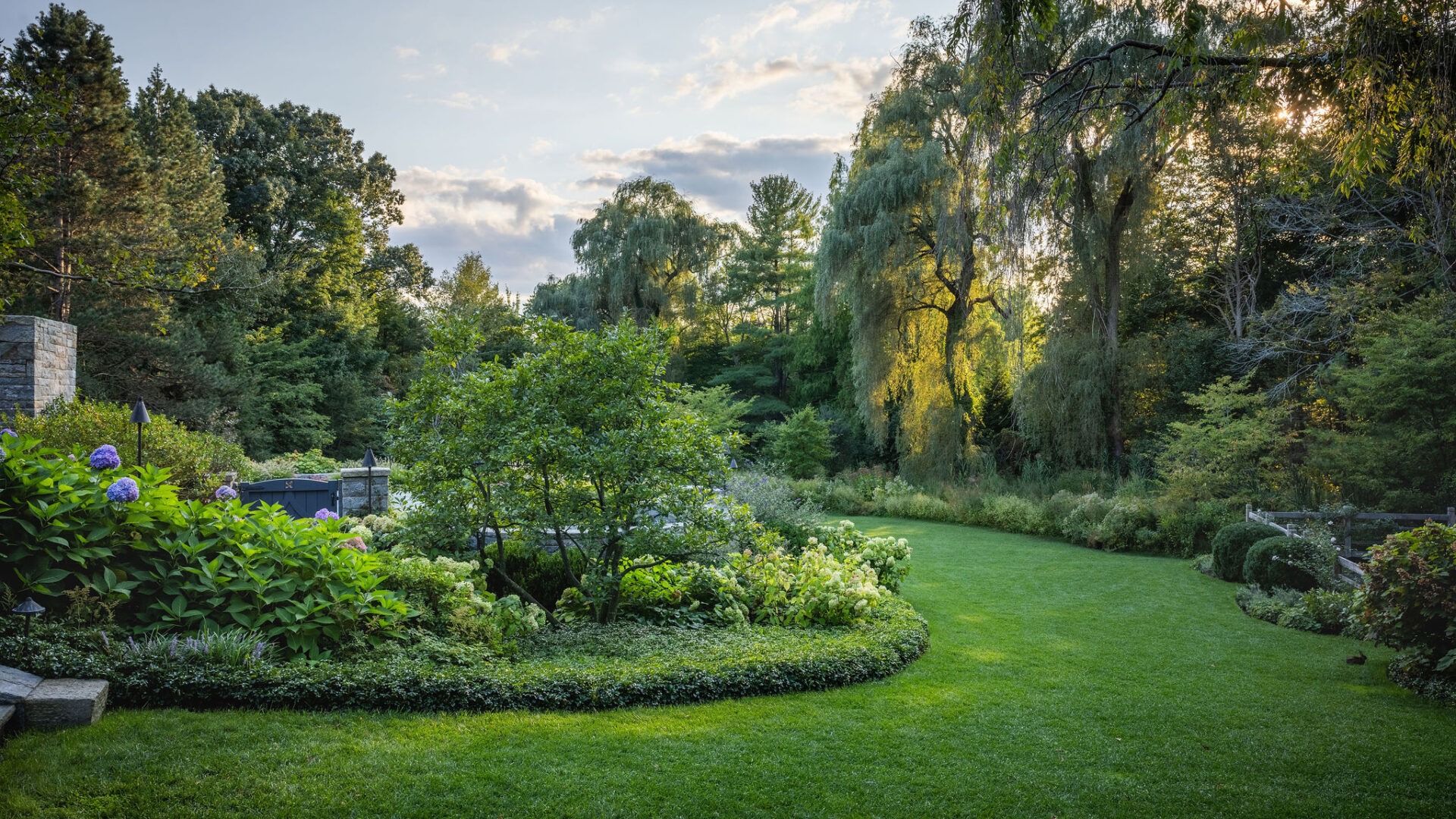 The image features a serene garden with lush green grass, trimmed hedges, flowering shrubs, and weeping trees, bathed in soft sunlight.