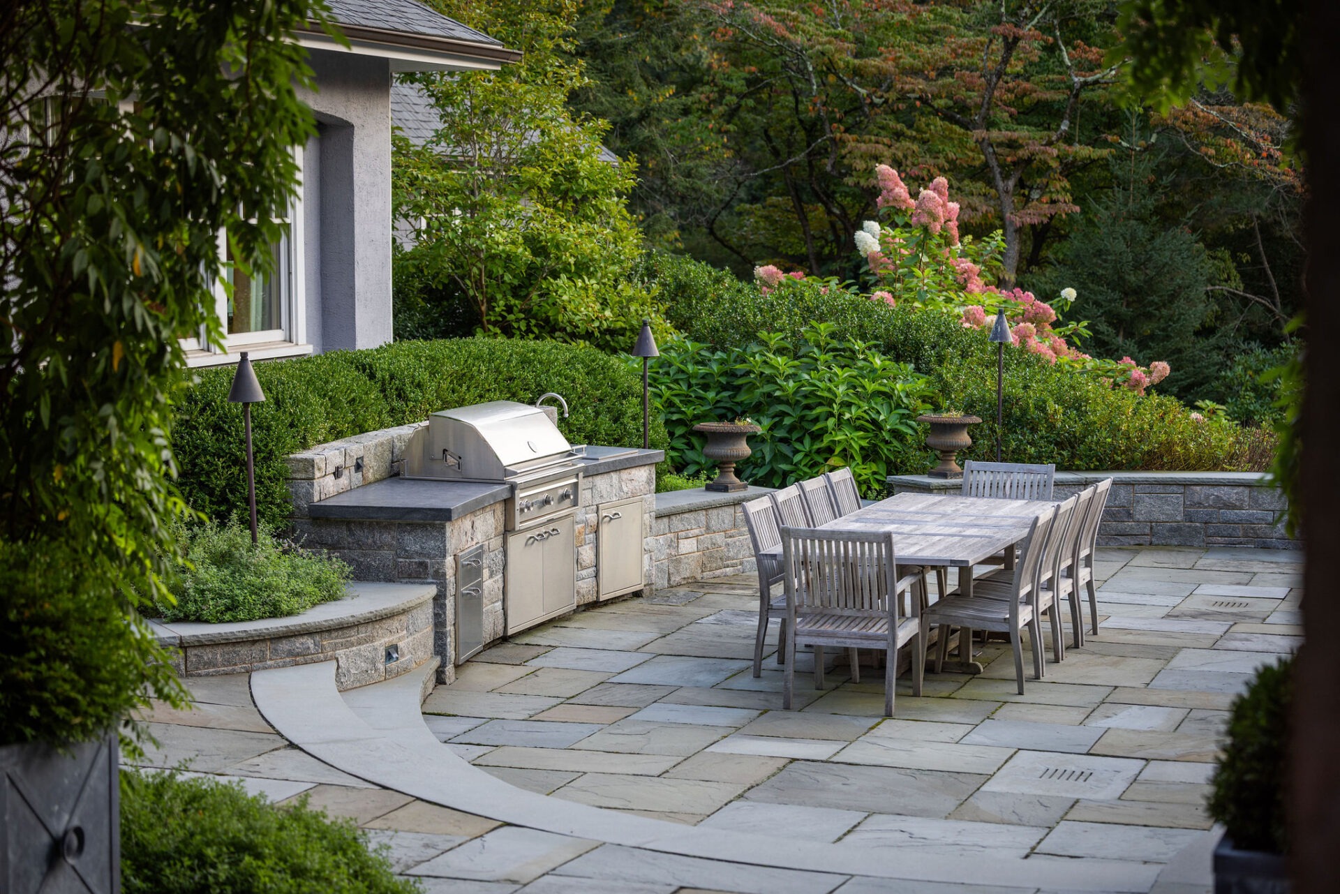 An outdoor stone patio with a built-in grill, a dining table with chairs, surrounded by lush greenery and flowering plants in a residential backyard.