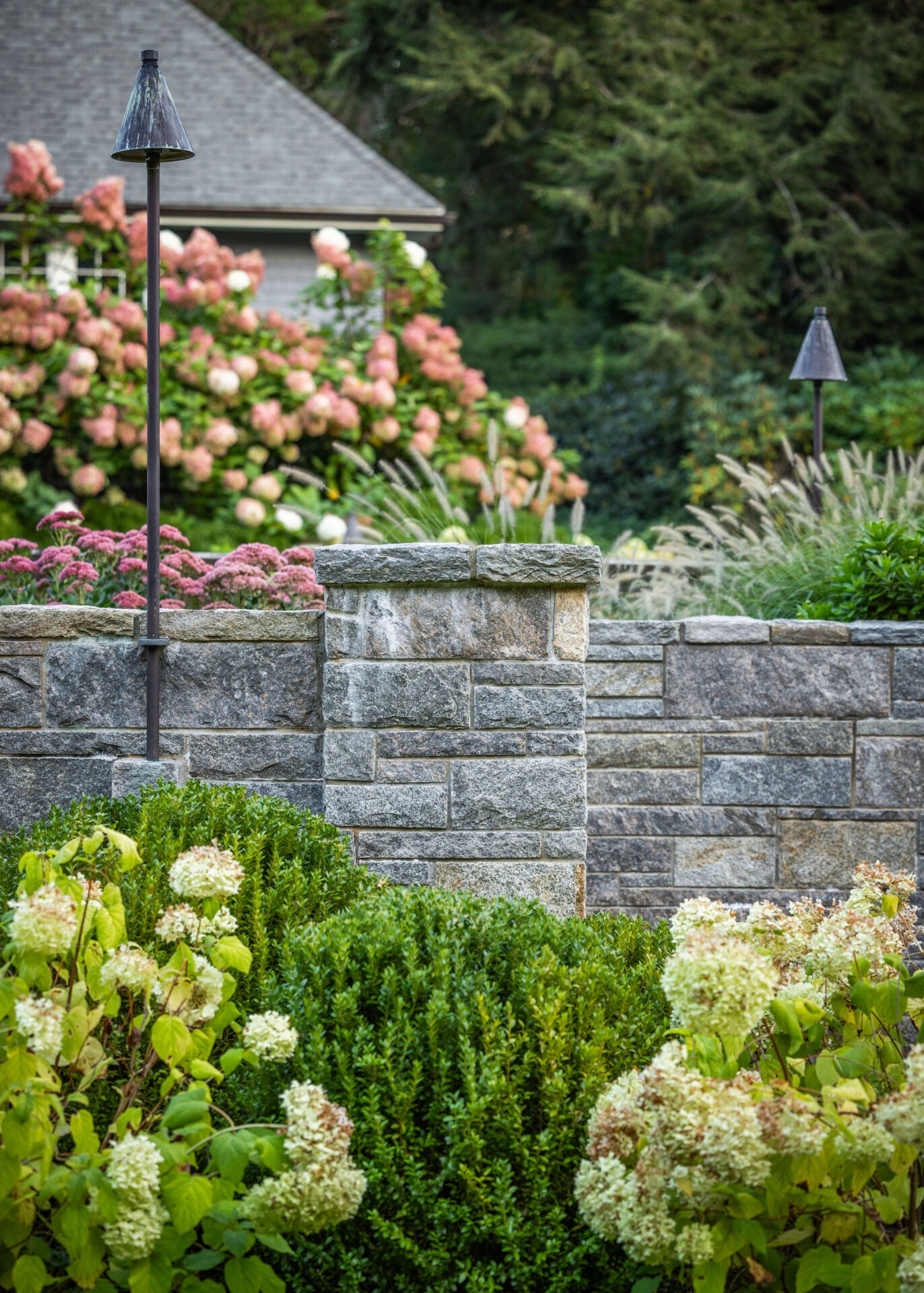A landscaped garden with a stone wall, lush greenery, hydrangea flowers, and a path lamp. A house and trees are visible in the background.