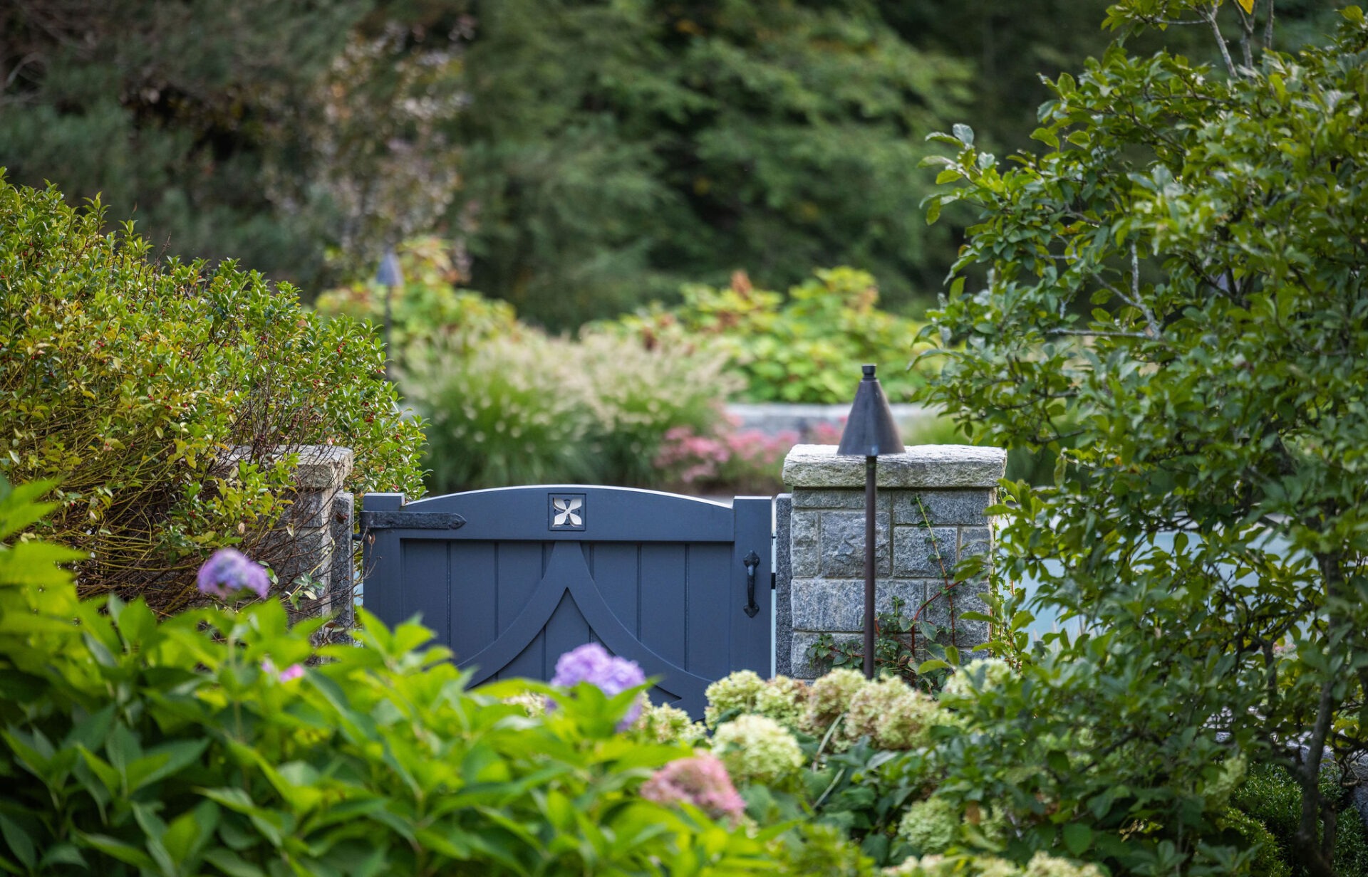 A blue gate with decorative hardware set in a stone wall, partially hidden by lush greenery and blooming flowers in a tranquil garden setting.