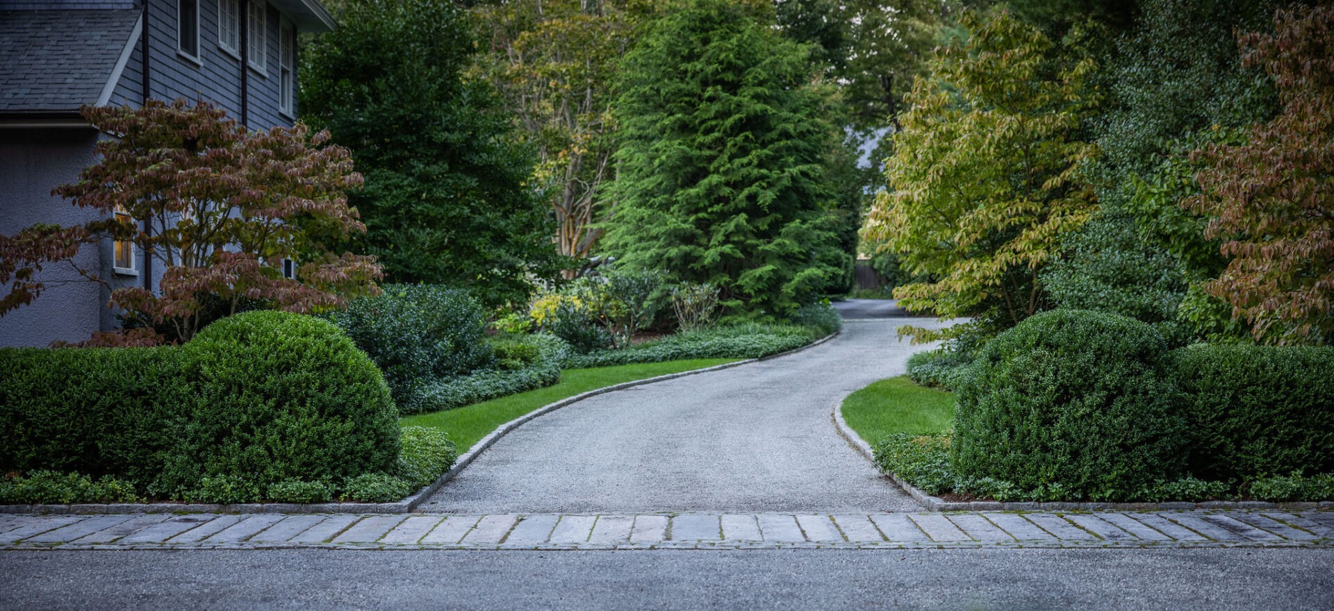 A winding cobblestone driveway leads through a well-manicured garden with trimmed hedges and trees beside a house's partial exterior.