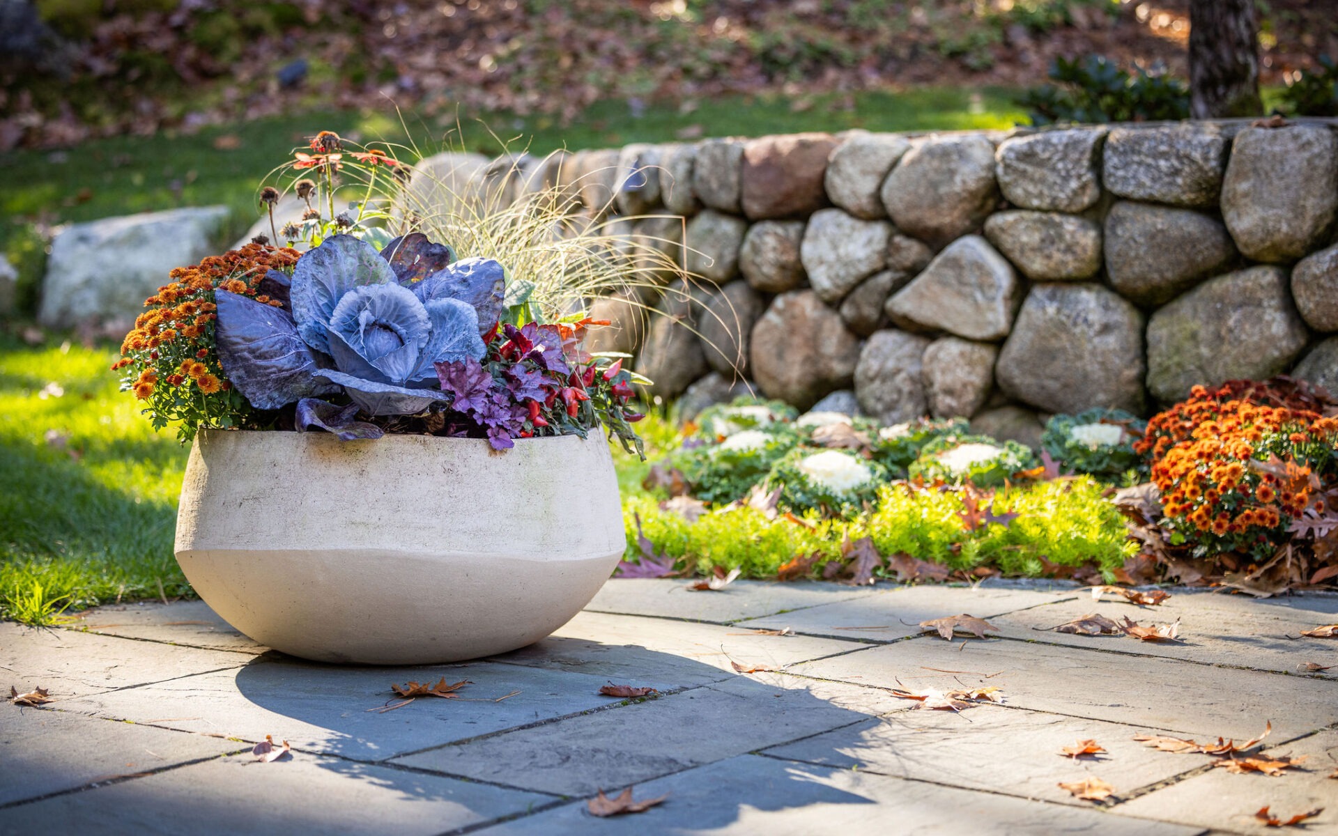 A large planter with blue and orange flowers sits on a stone-paved patio with a stone wall and autumn leaves scattered around.