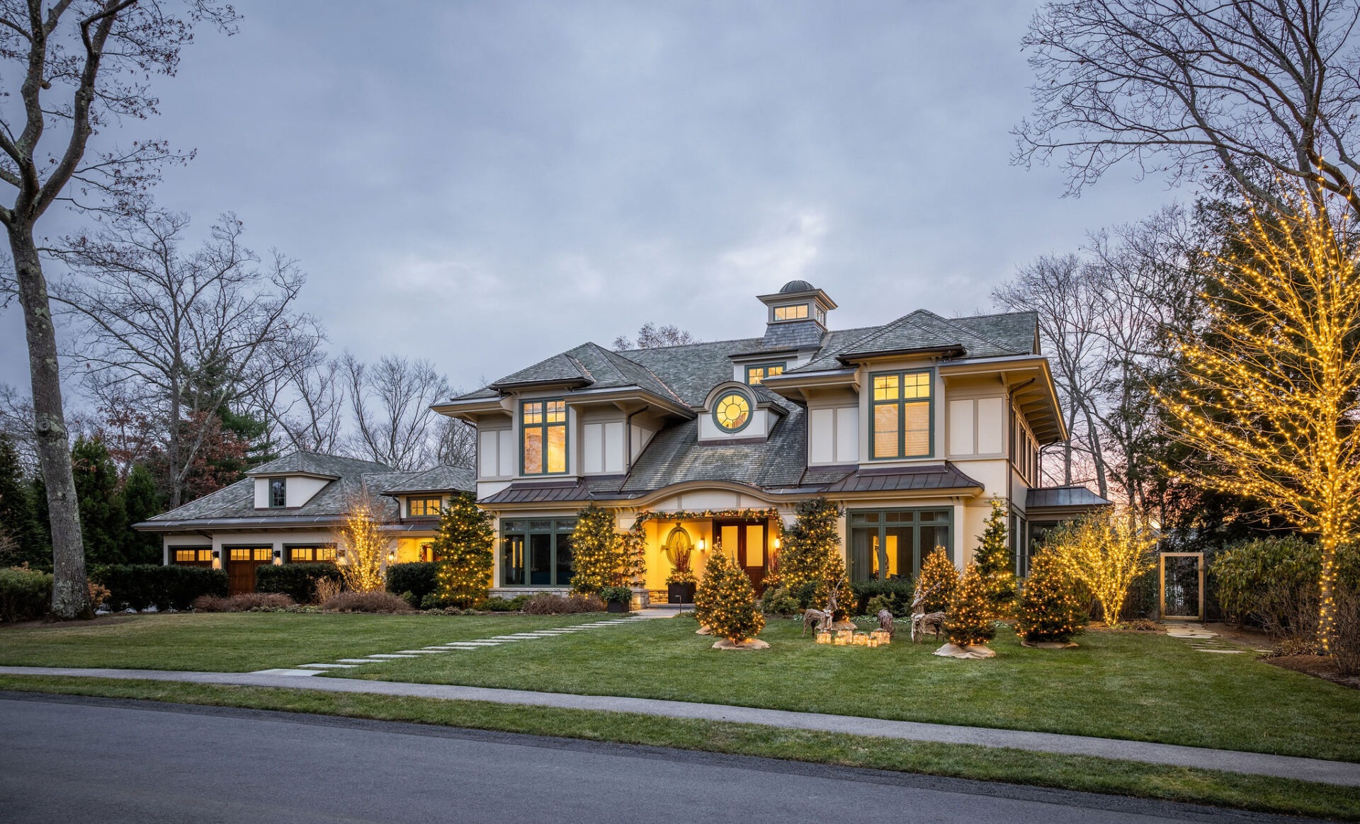 A large, elegant two-story house with a well-manicured lawn, warm exterior lighting, and holiday decorations under a twilight sky.