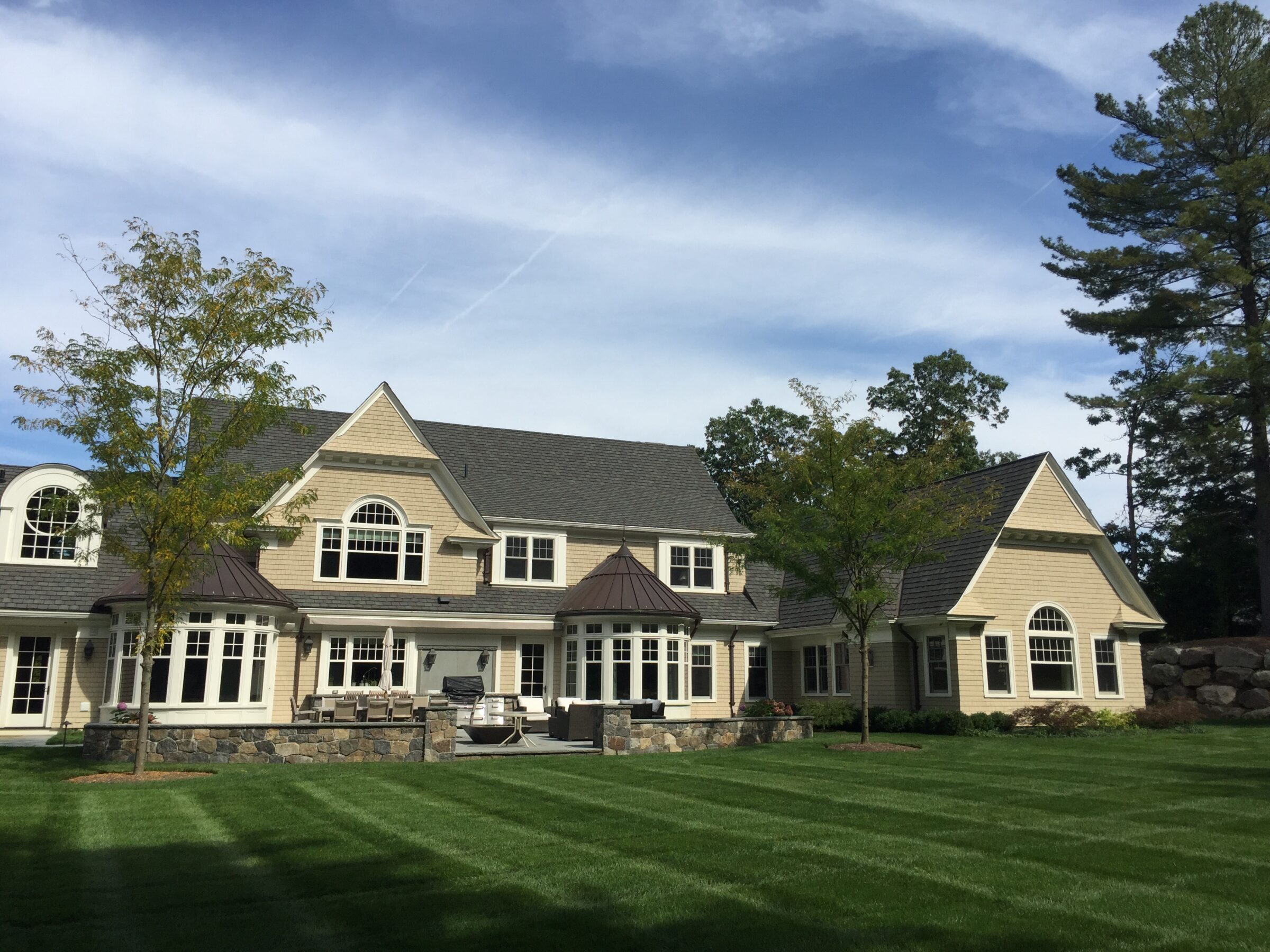 A large suburban house with beige siding, stone accents, multiple gables, and arched windows, surrounded by a manicured lawn and trees under a blue sky.