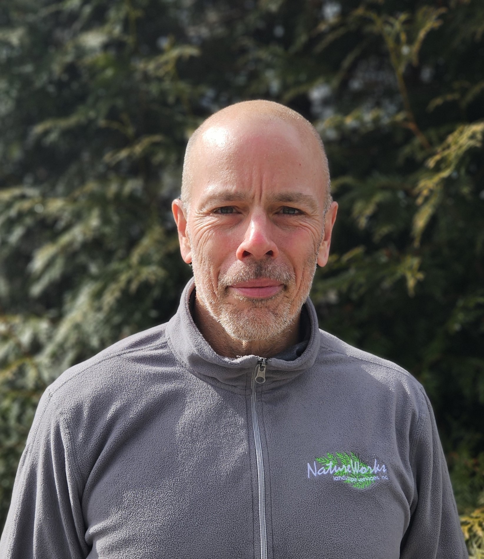 A person with a bald head and goatee is wearing a grey fleece jacket outdoors, with greenery in the background, partially smiling at the camera.