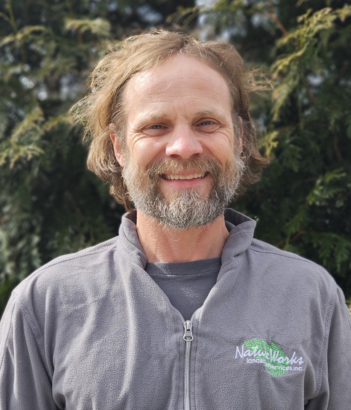 A smiling person with a beard and mustache, wearing a gray fleece jacket, stands in front of a tree with green foliage.