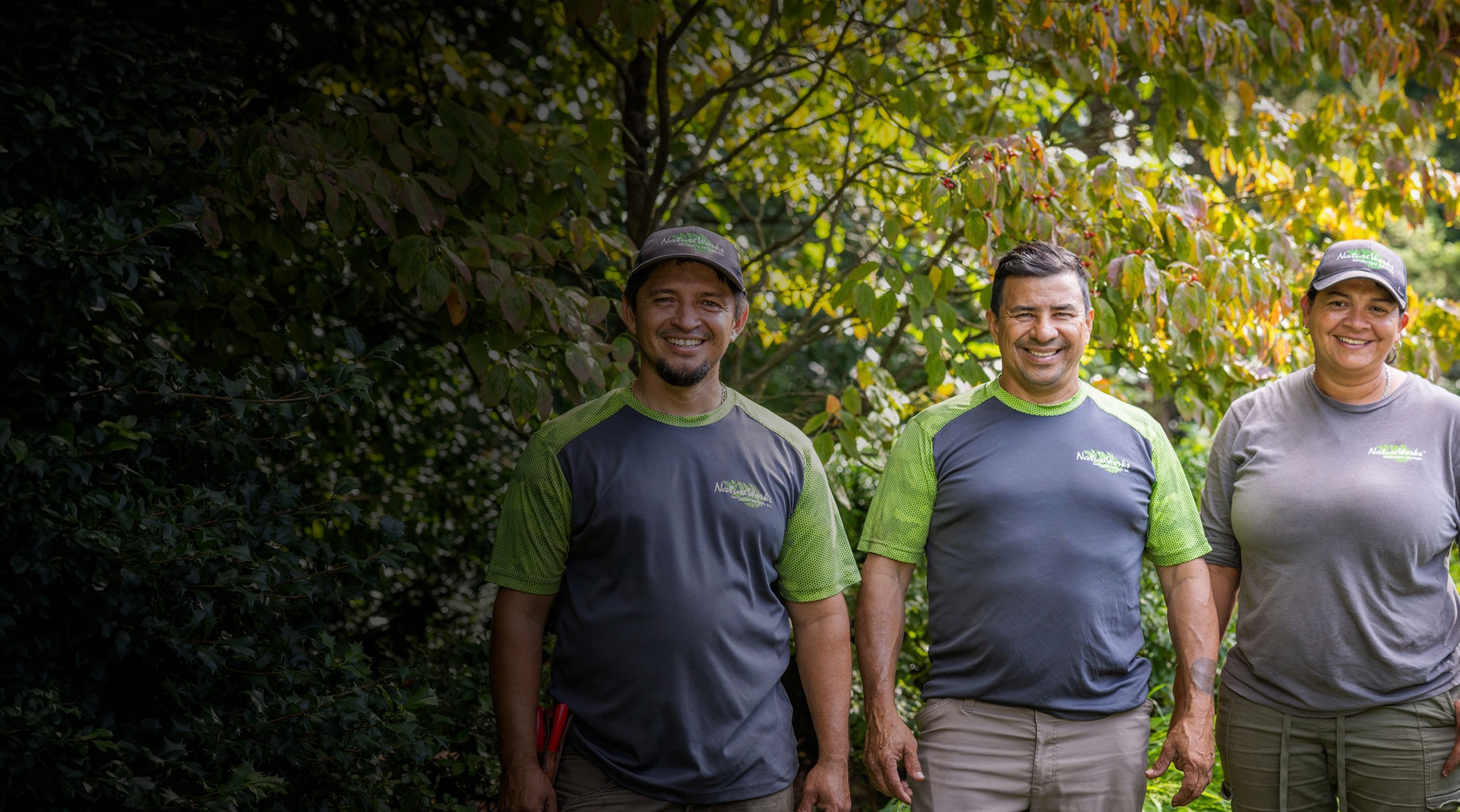 Three smiling people in matching green and gray uniforms stand in front of lush green foliage, possibly a team or coworkers outdoors.
