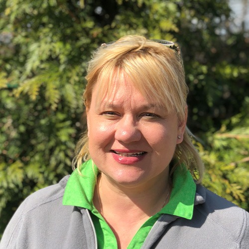 This image shows a smiling person with blonde hair outdoors on a sunny day, wearing a gray and green shirt, with greenery in the background.