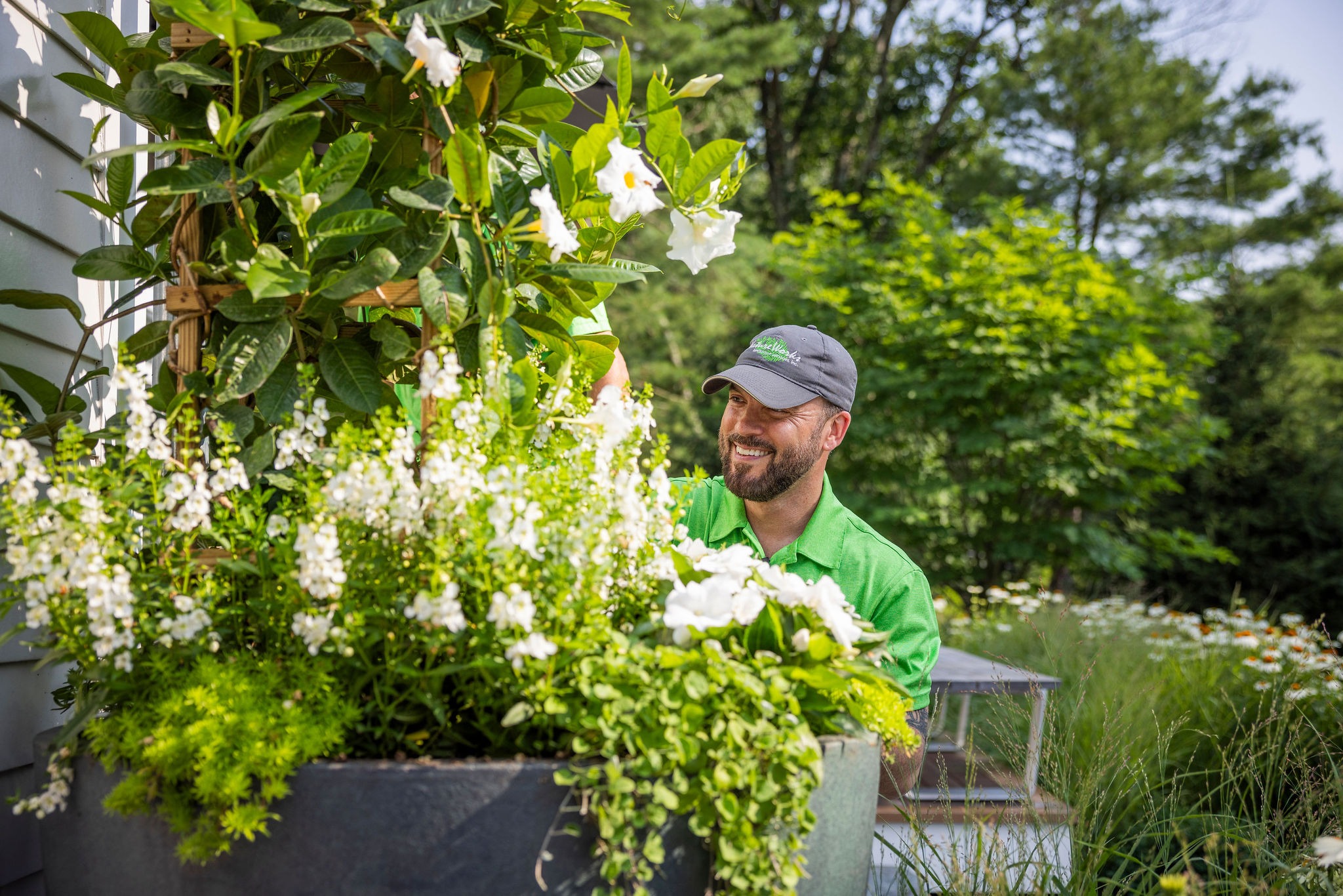 A person in a green shirt and cap smiles while tending to lush, flowering plants in a sunlit garden with trees in the background.