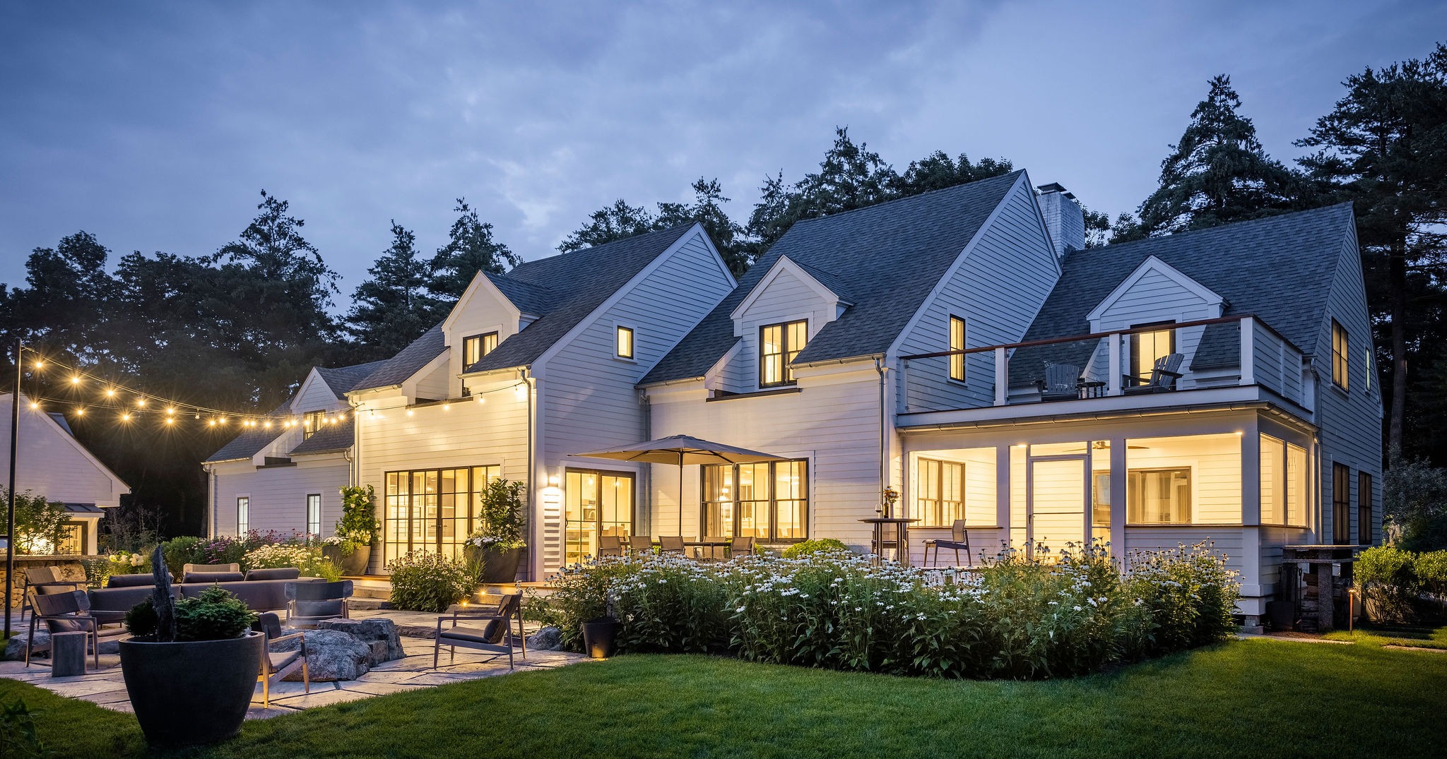 This image captures a large, elegant house at dusk with outdoor lighting, comfortable patio furniture, landscaped garden, and string lights adding ambiance.