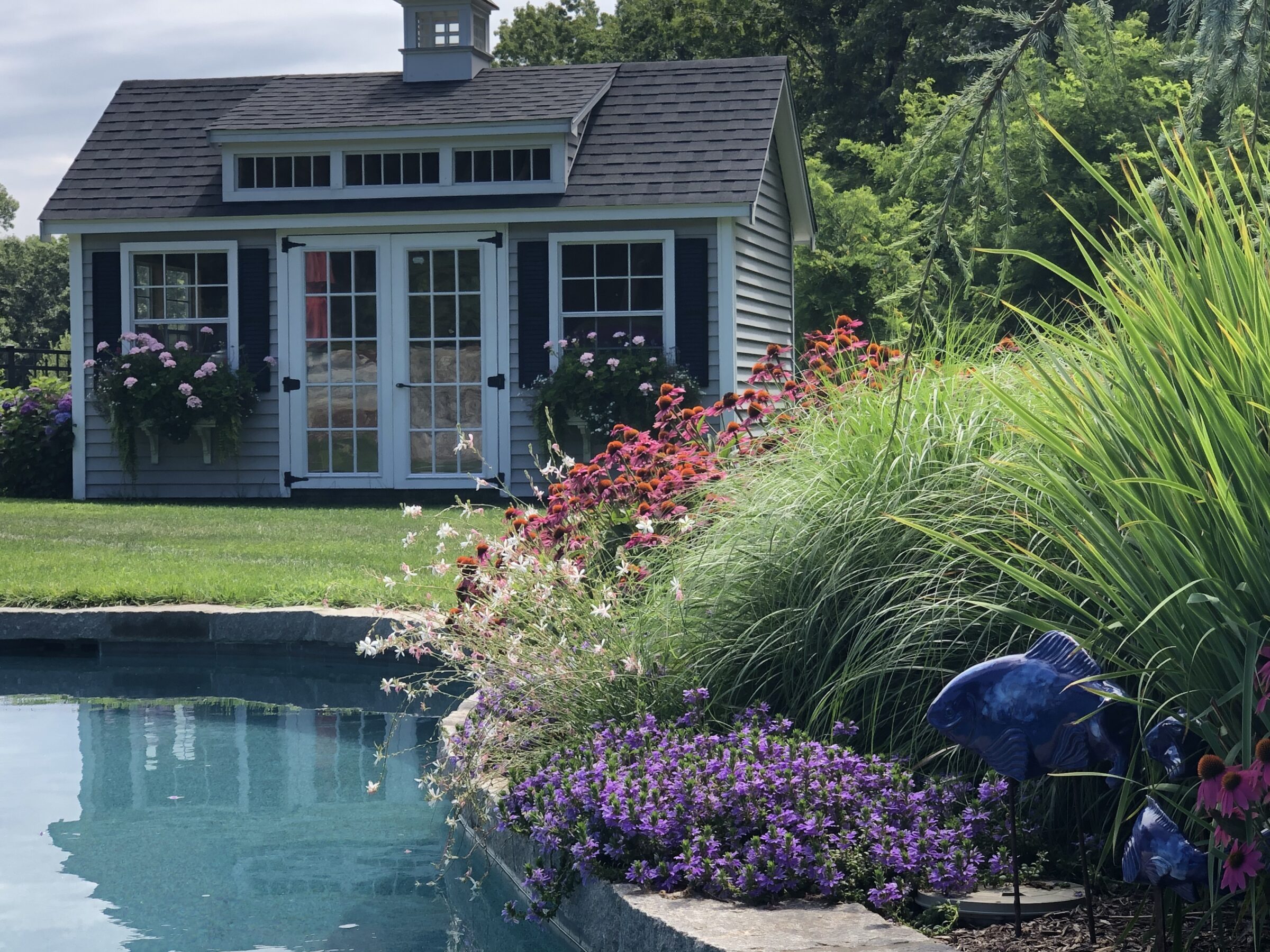 A quaint garden shed with French doors surrounded by lush flowers overlooks a serene pool with ornamental grasses and artistic fish sculptures.