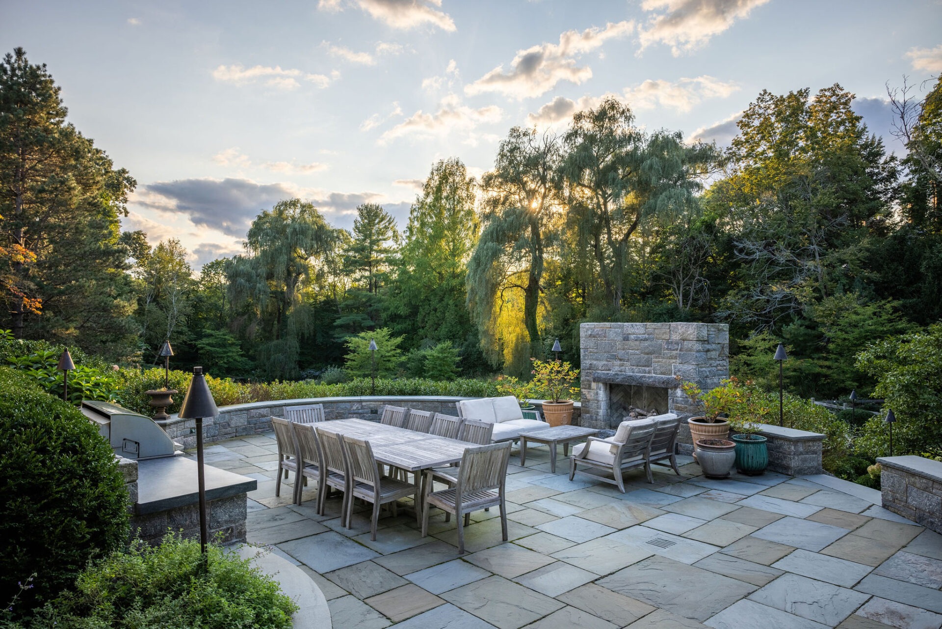 An elegant outdoor patio with a dining table, chairs, a lounging area, and a stone fireplace, set against a backdrop of lush trees and sunset.
