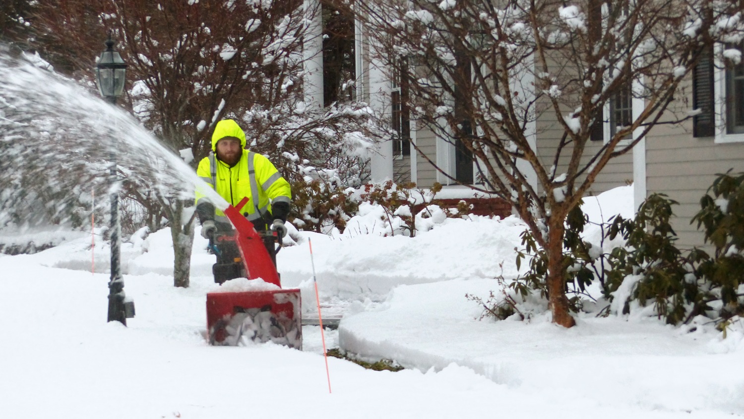 A person in a high-visibility jacket operates a red snowblower, clearing snow in front of a residential house with bare trees and shrubs around.