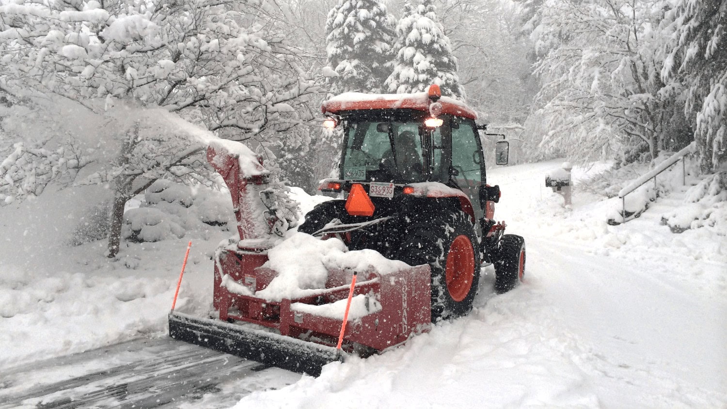 A snow-covered landscape with heavy snowfall. A person operates an orange snowblower tractor clearing a path on a residential street. Trees line the area.