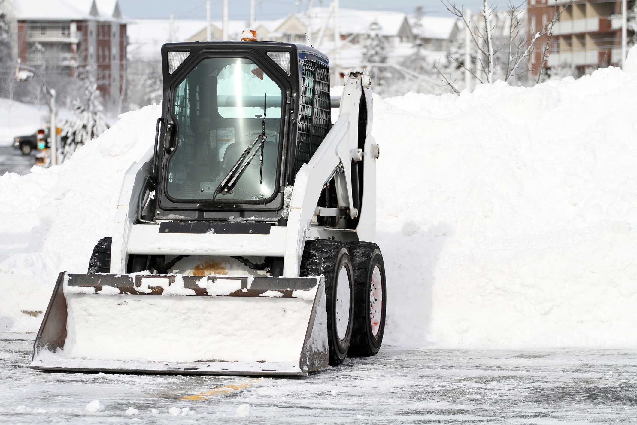 A skid-steer loader with a snow bucket attachment is positioned near a large pile of snow in a cleared parking lot, suggesting recent snow removal efforts.