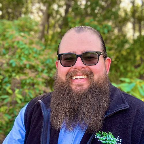 A smiling person with a long beard, wearing sunglasses and a vest, stands before a blurred background of green foliage outdoors.